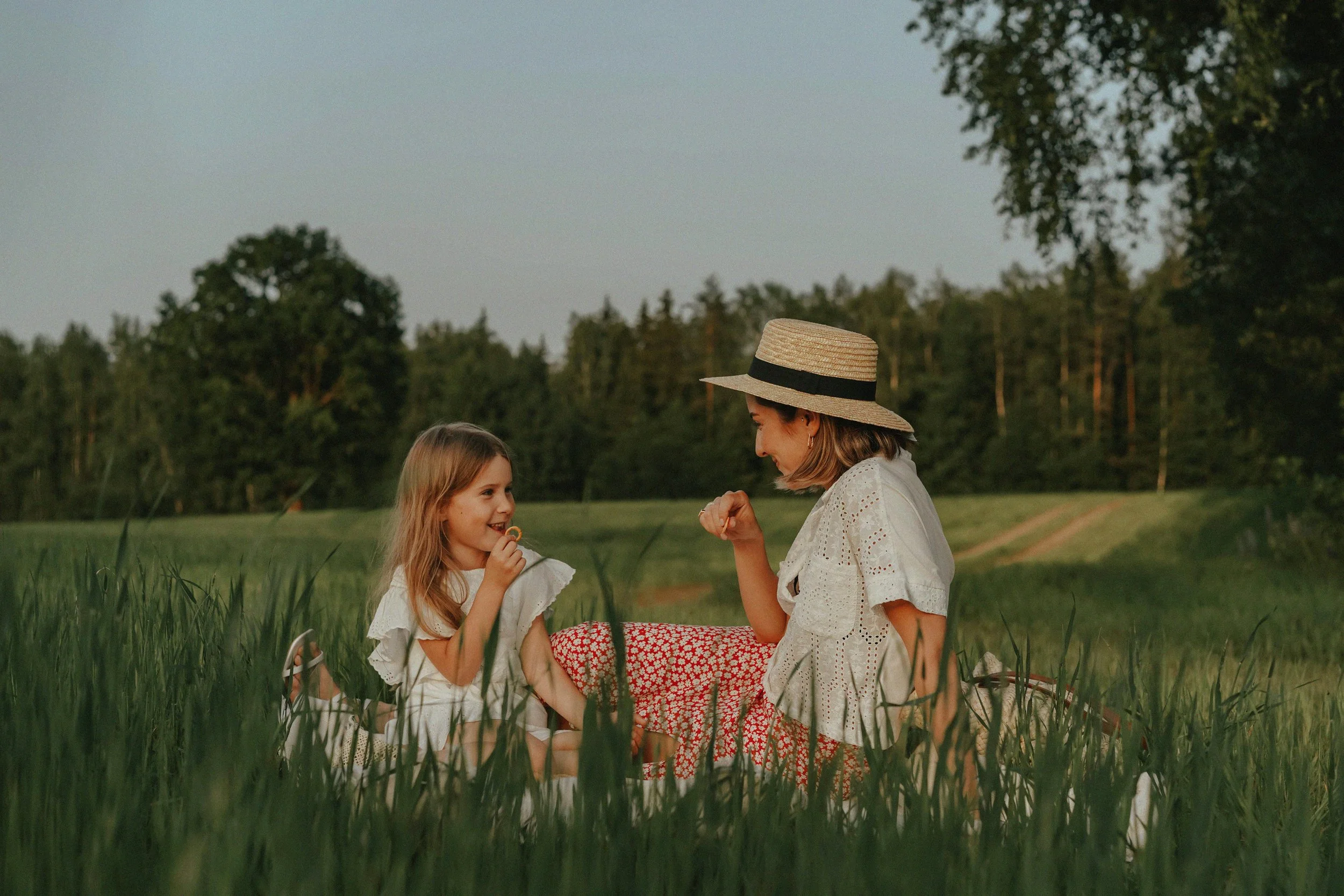 A woman and a young girl sitting in tall grass in a field, smiling and playing a game together, with trees in the background and a clear sky overhead.