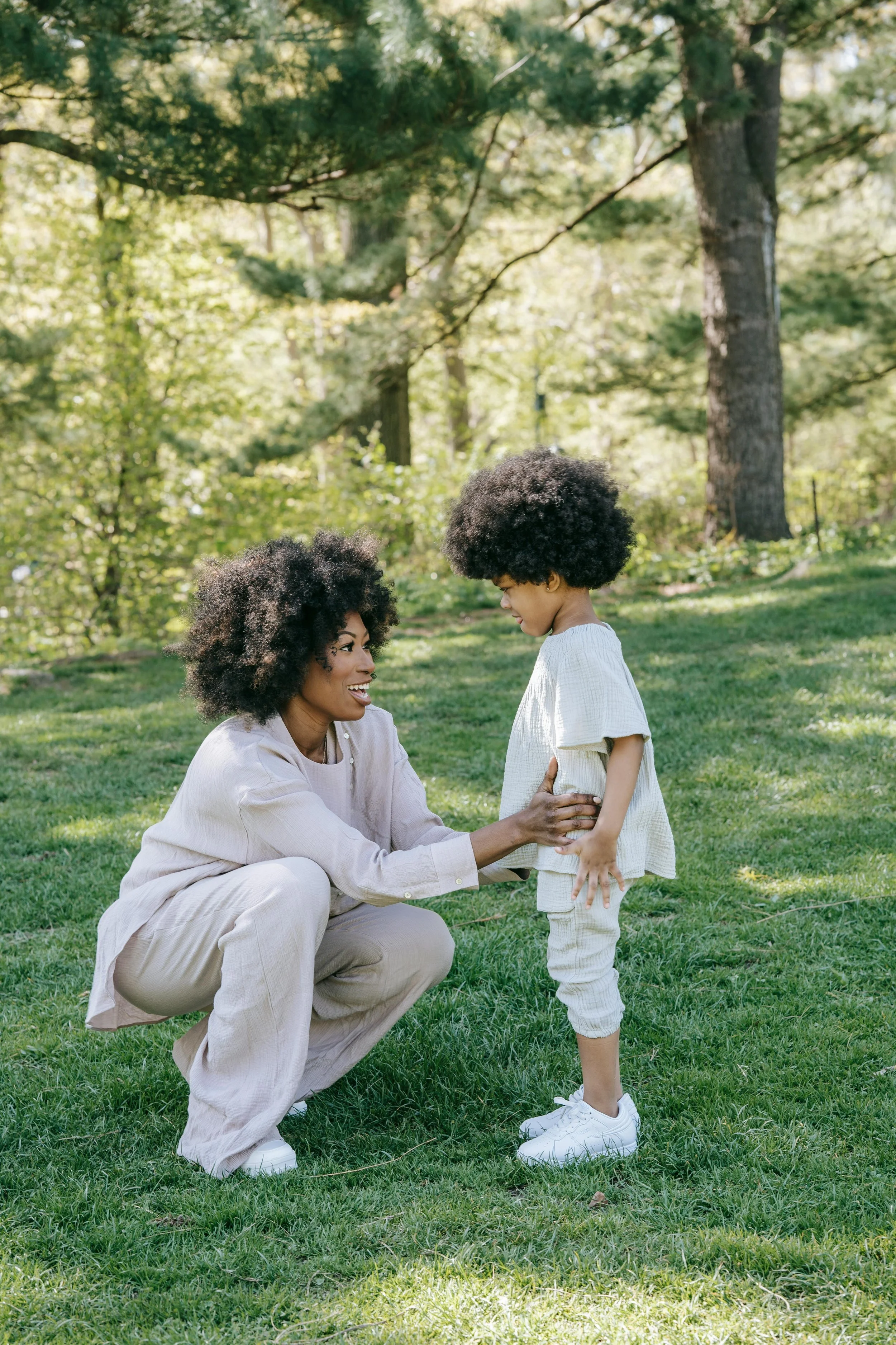 A woman and a young girl with curly hair are talking and smiling in a park with green grass and trees.