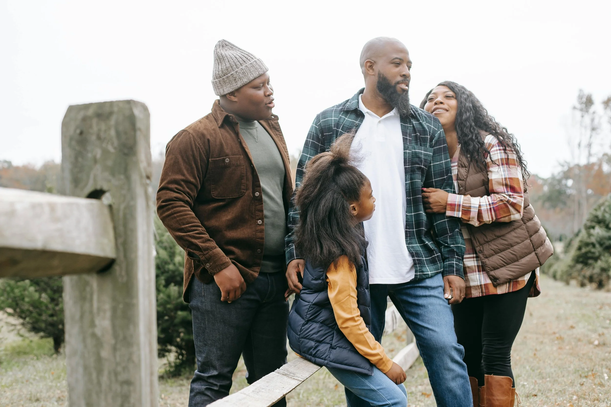 Family of five standing outdoors near a wooden fence with trees and cloudy sky in the background