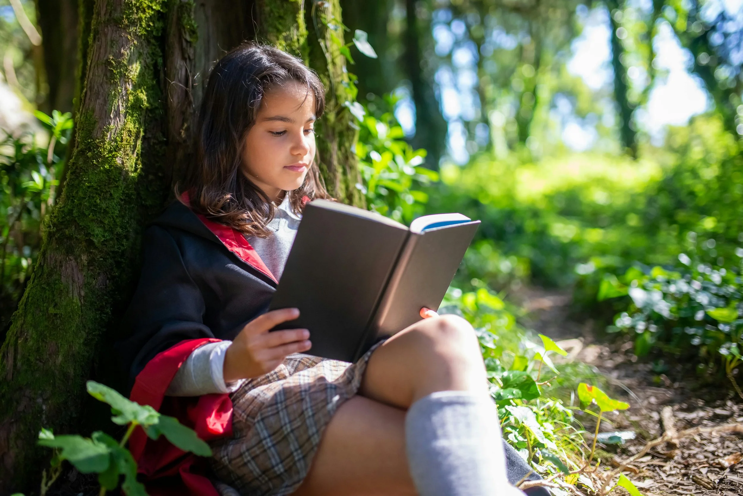 Girl sitting against a tree in a forest, reading a book