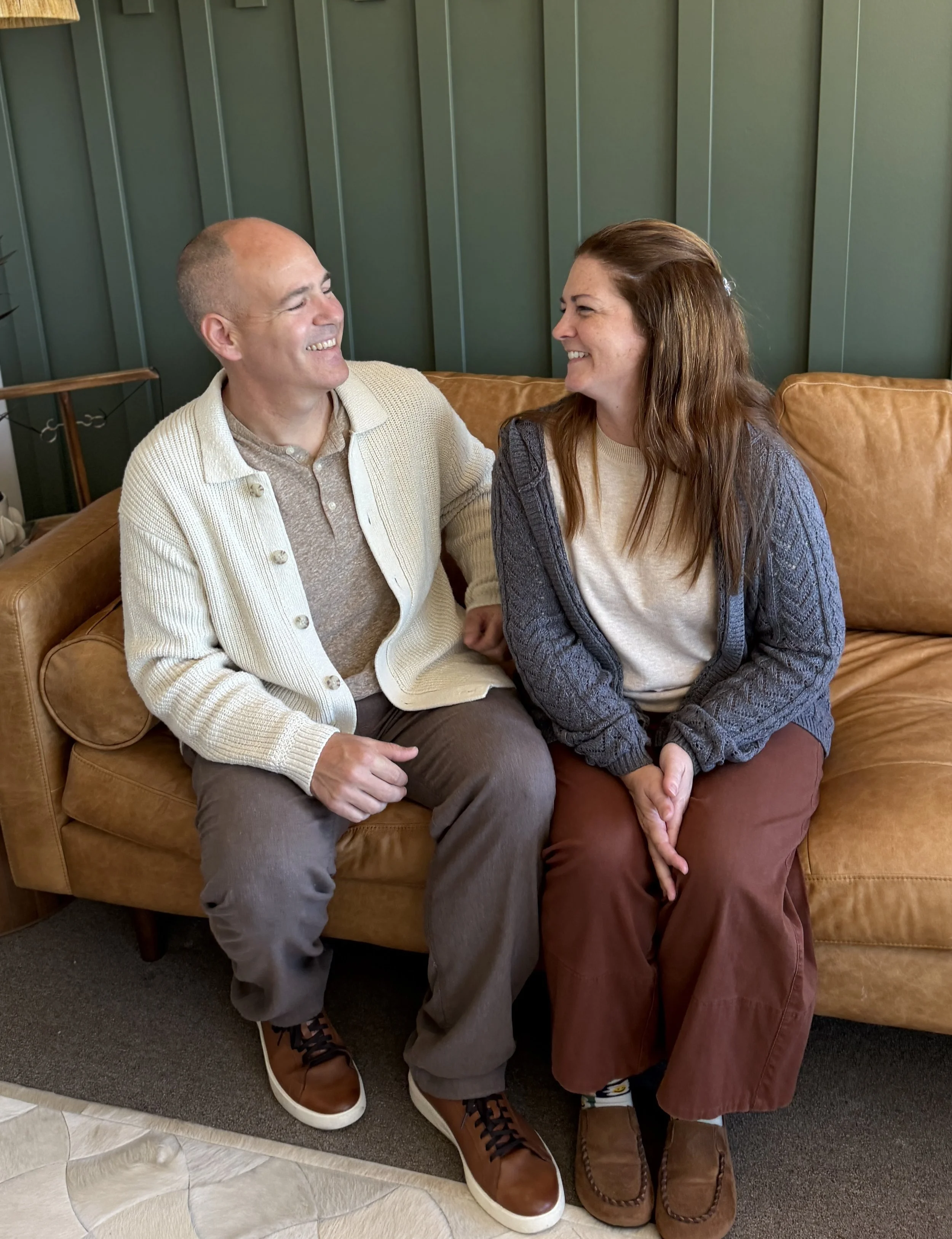 A man and a woman sitting close together on a brown leather couch, smiling at each other, in a room with green panel walls.