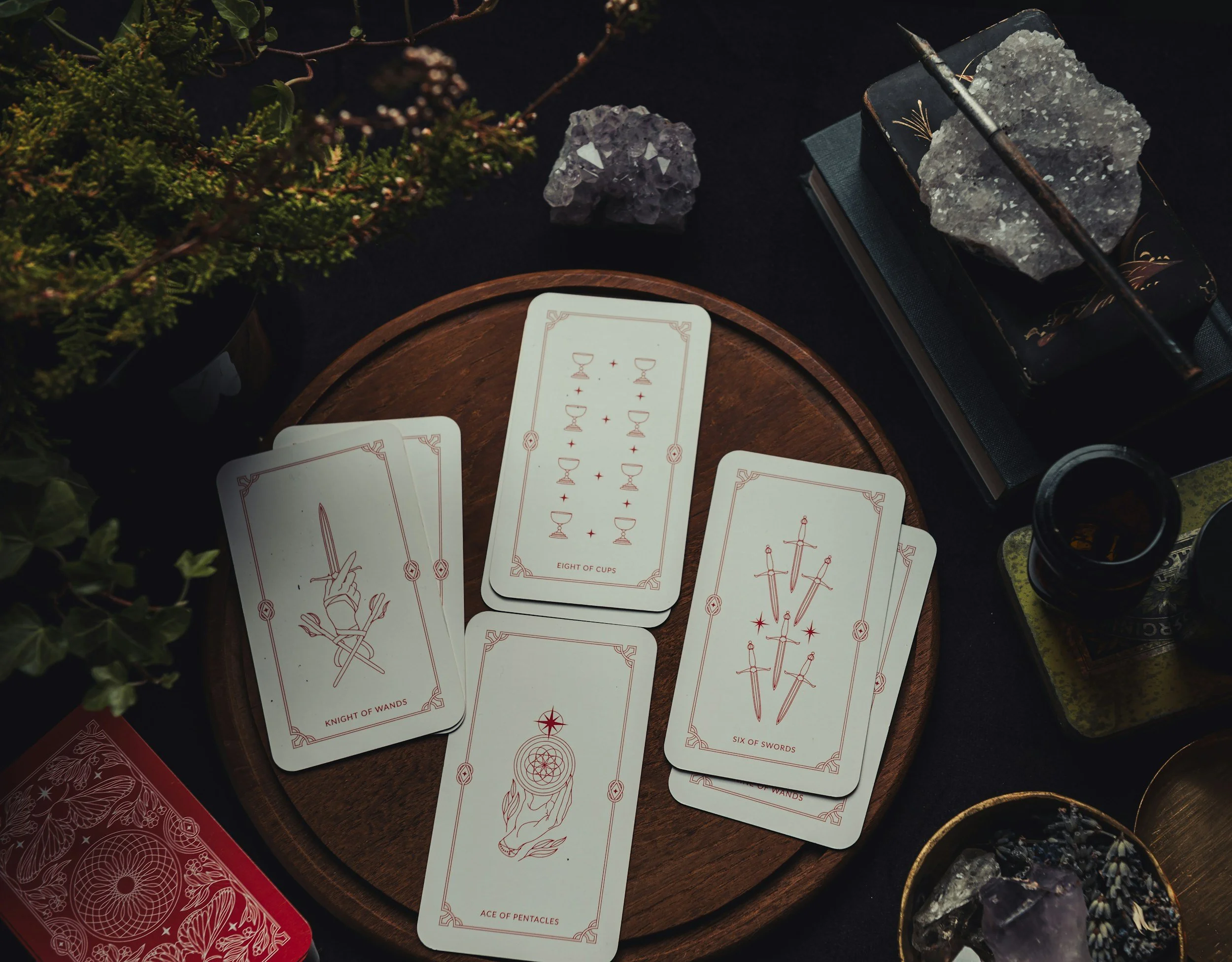Tarot cards arranged on a wooden plate with various crystals and a black cup, surrounded by greenery and books.