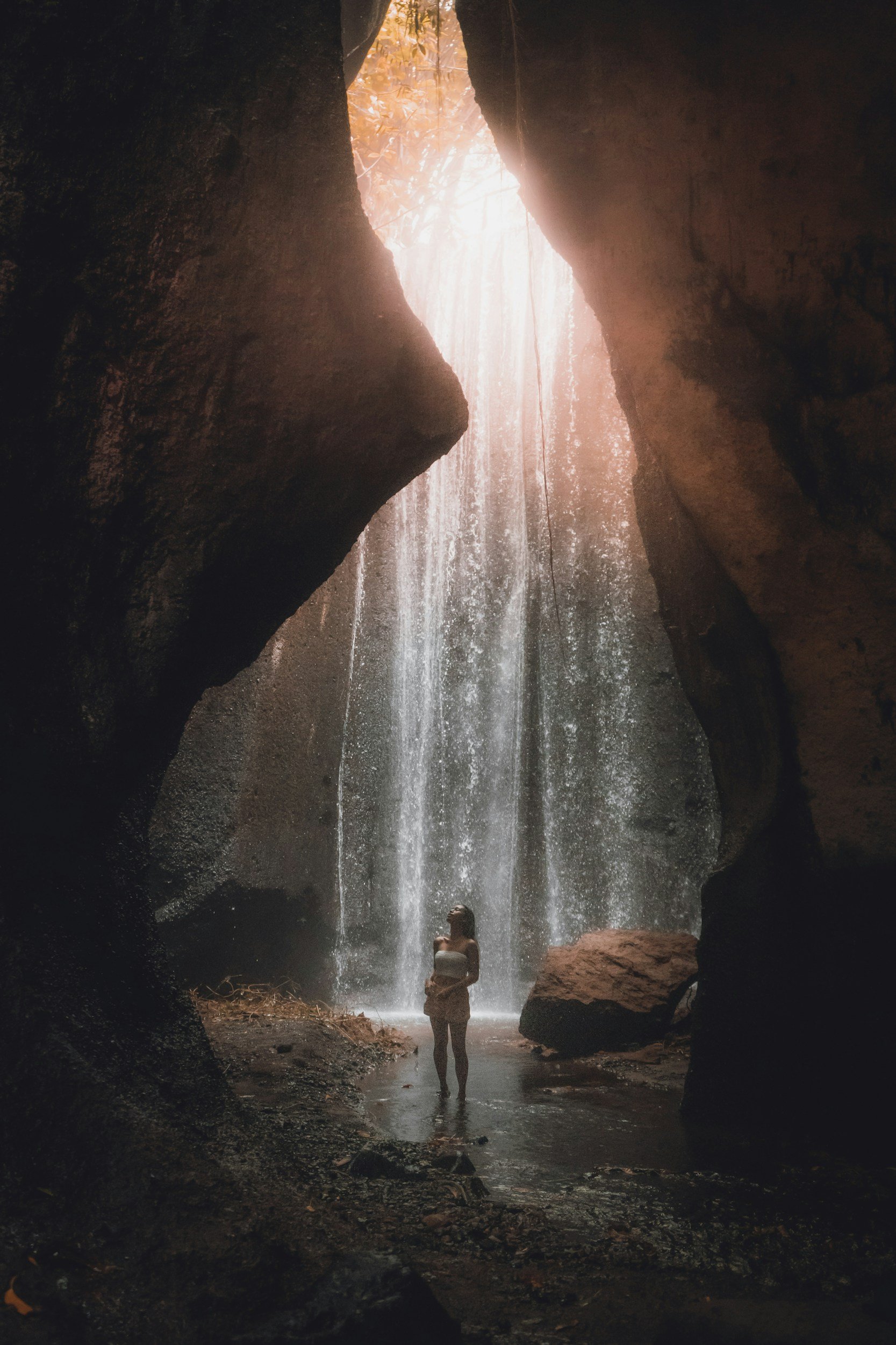 A person standing in front of a waterfall inside a cave with sunlight filtering through.