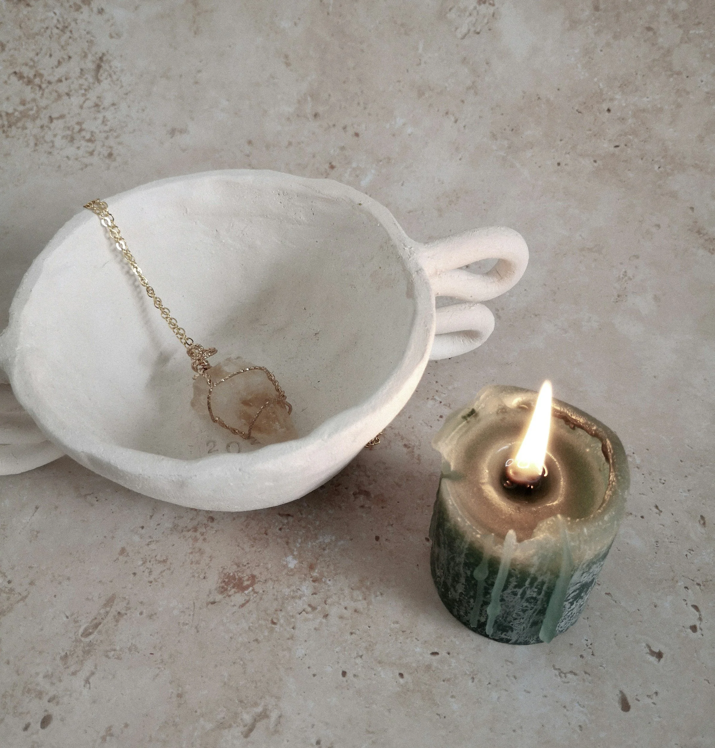 A white ceramic cup with extended handles holds a gold necklace with a pendant; a lit green candle with white wax drips sits nearby on a beige surface.