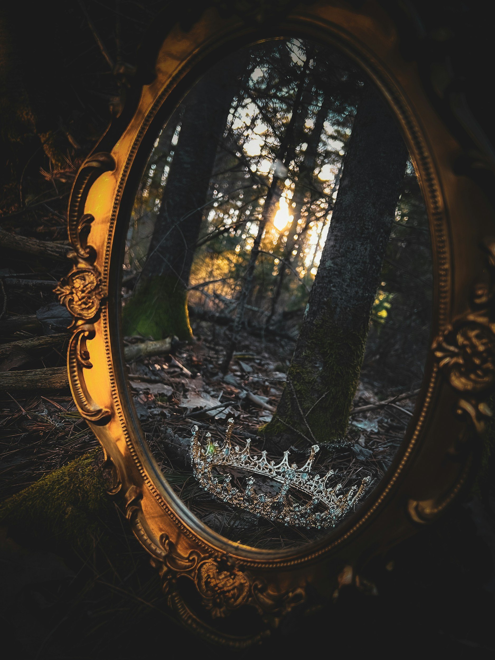 A decorative mirror on the forest floor reflecting trees and sunlight, with a tiara beside it.