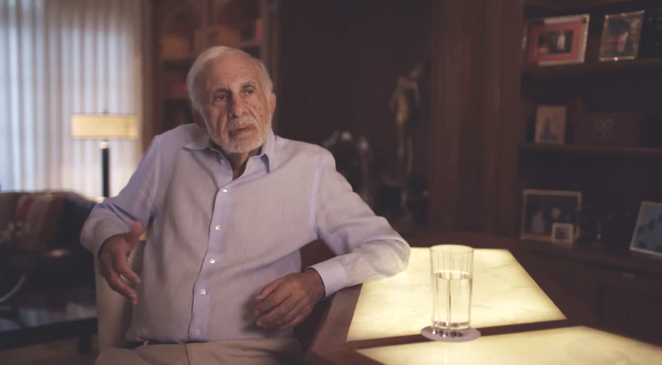 Carl Icahn wearing a white button-up shirt, sitting at a wooden desk with a glass of water, in a warmly lit room with framed photos and decorations in the background.
