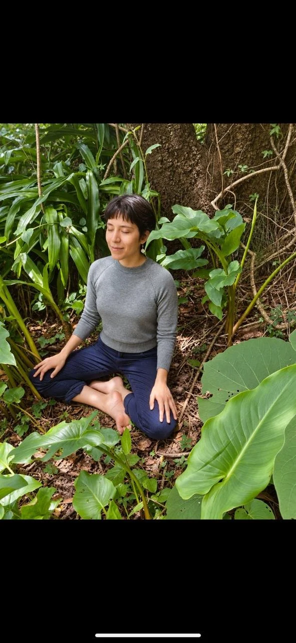 Mujer con camisa gris y pantalones oscuros, sentada en posición de meditación en un entorno selvático rodeada de plantas verdes grandes y árboles. Baño de bosque