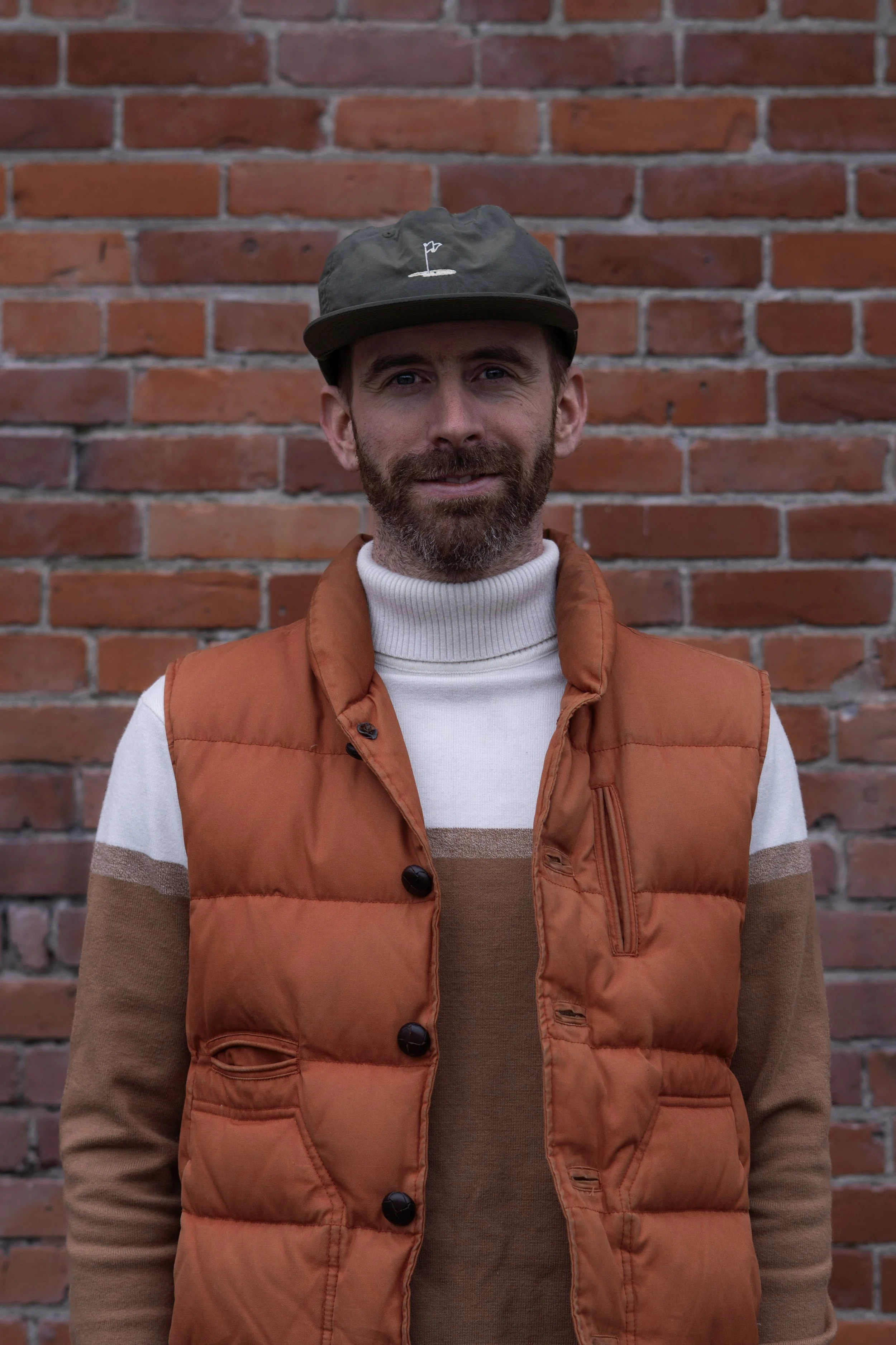 Portrait of a man with a beard and short hair, wearing a gray sweatshirt over a dark collared shirt, standing in front of a brick wall.