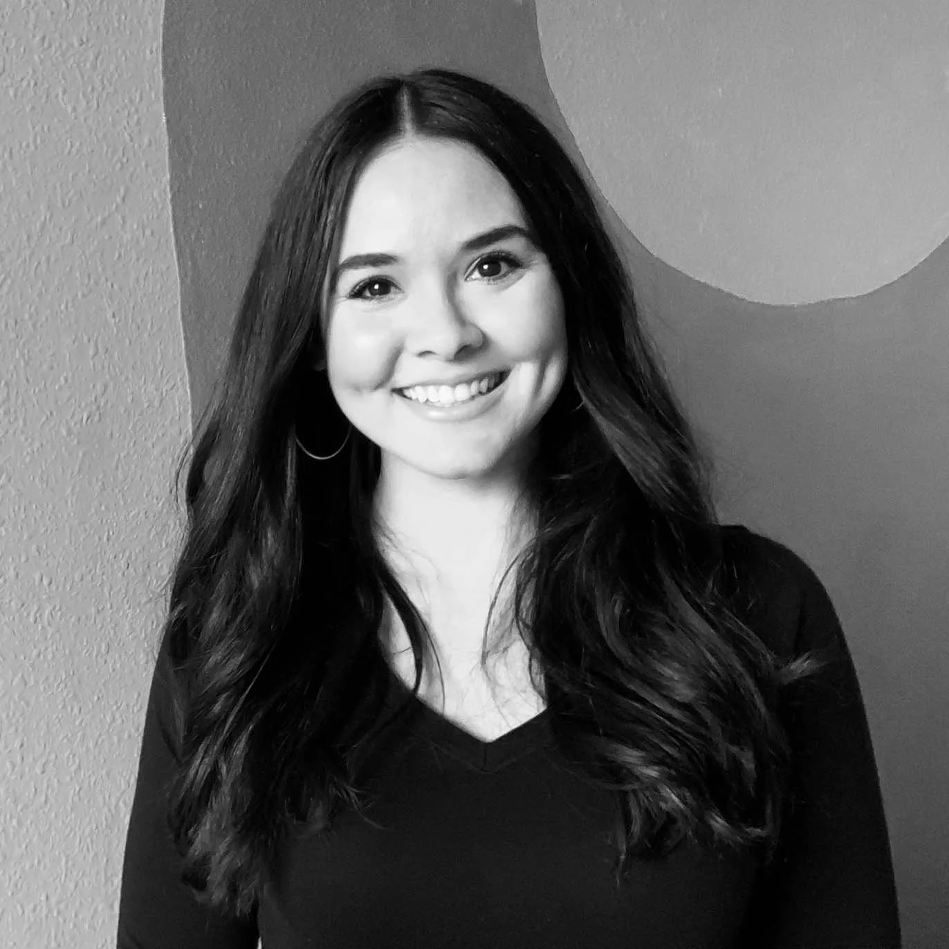 A young woman with long dark wavy hair smiling and wearing hoop earrings, standing in front of a two-tone wall with a circle design.