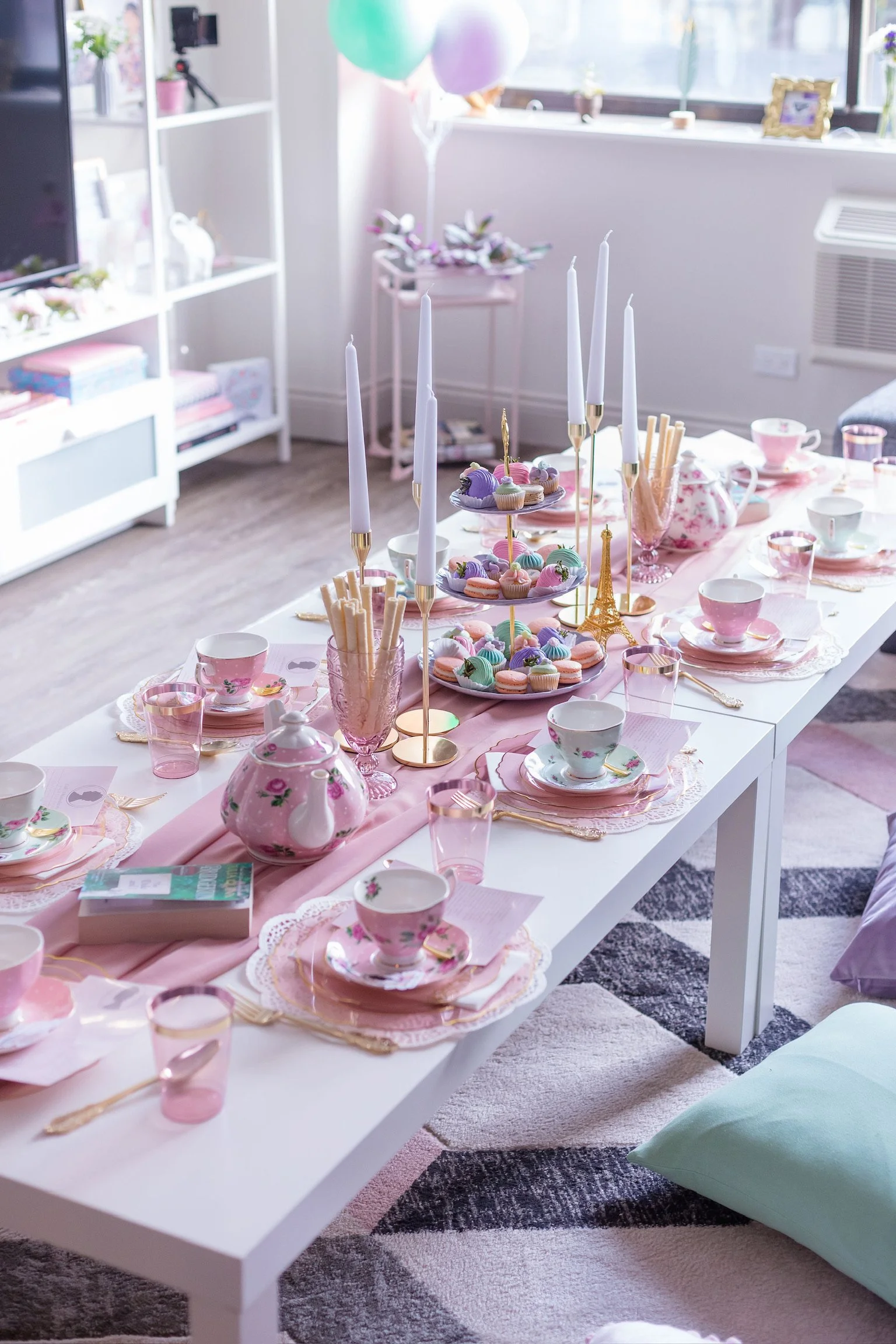 A decorated dining table with pink tablecloth for a tea party, featuring pastel-colored teacups, plates, and a variety of desserts, including macarons and cupcakes, surrounded by pink glasses, gold cutlery, and centerpieces with tall white candles and a small gold Eiffel Tower ornament.