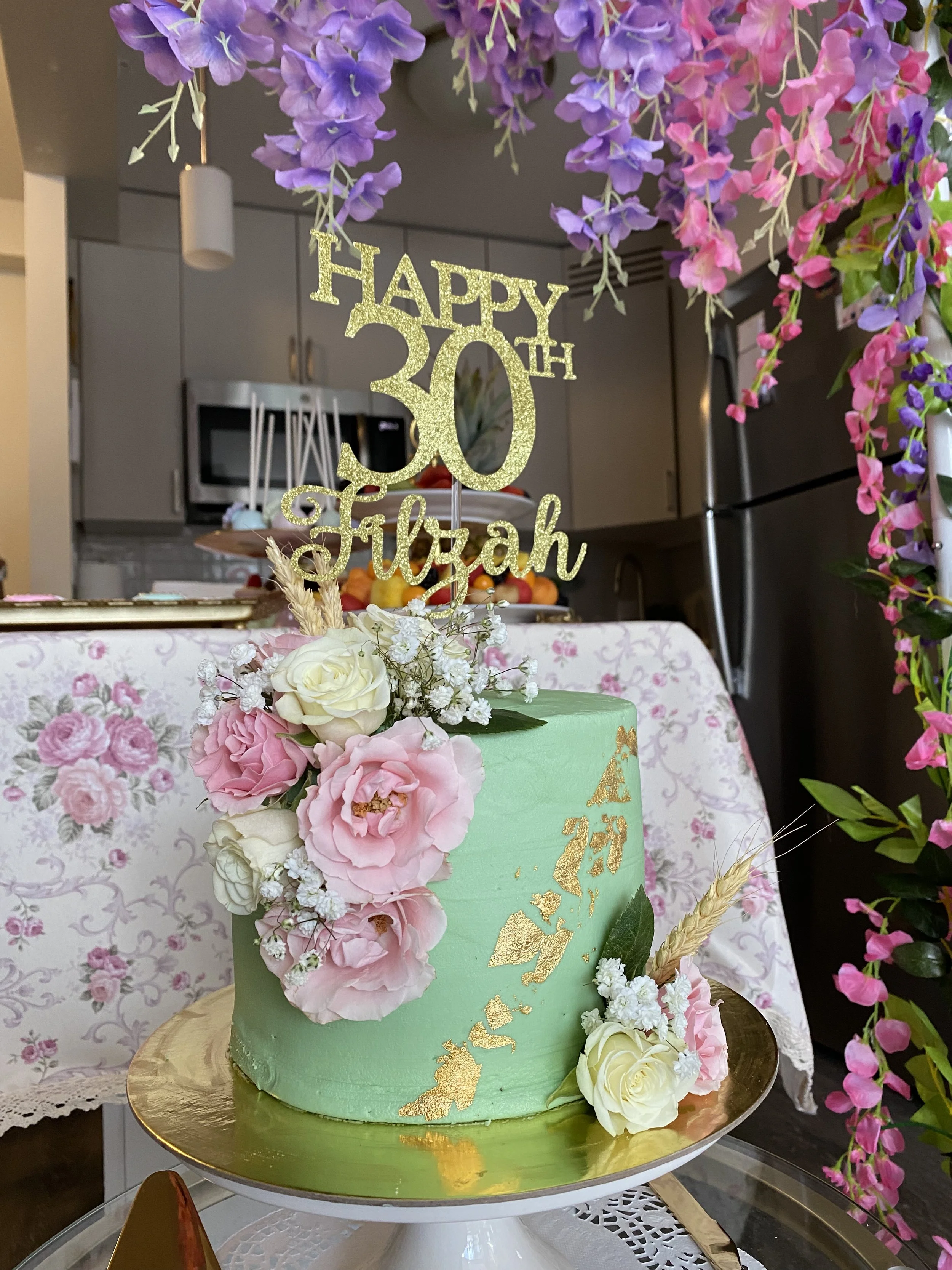 A two-tiered green birthday cake decorated with white and pink roses, white baby's breath, and gold foil accents, topped with a glittery gold 'Happy 30th' sign, surrounded by pink and purple flowers, on a white cake stand.