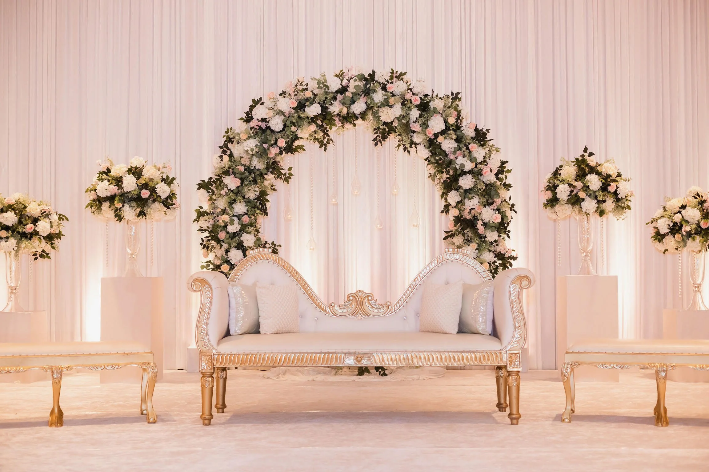 Elegant wedding stage setup with a white and gold sofa, floral arch, and flower arrangements.