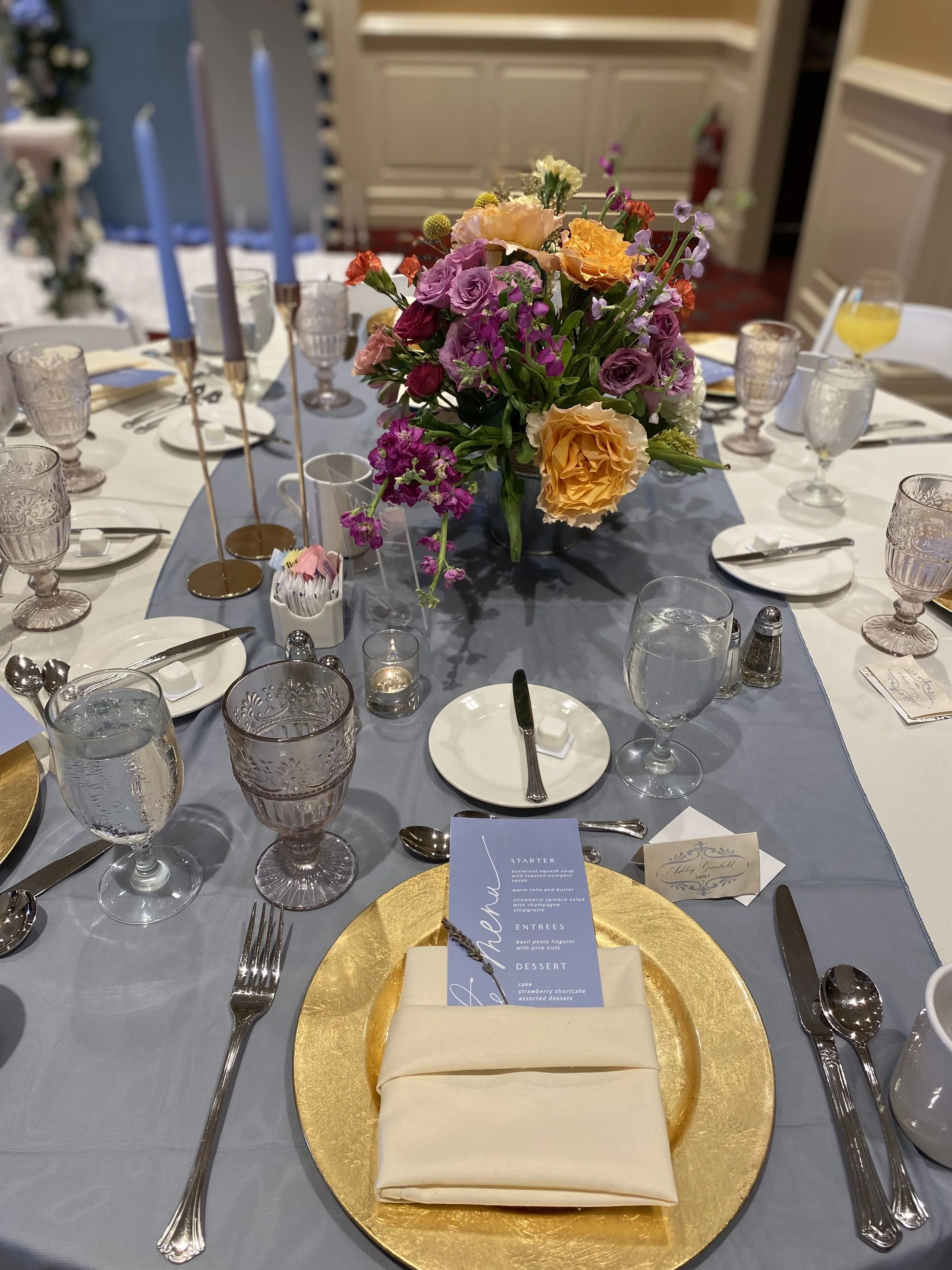 A table set for a formal dinner with a large bouquet of colorful flowers as the centerpiece, surrounded by wine glasses, water glasses, plates, silverware, and tall candles. A menu card is placed at the gold charger with a napkin underneath.