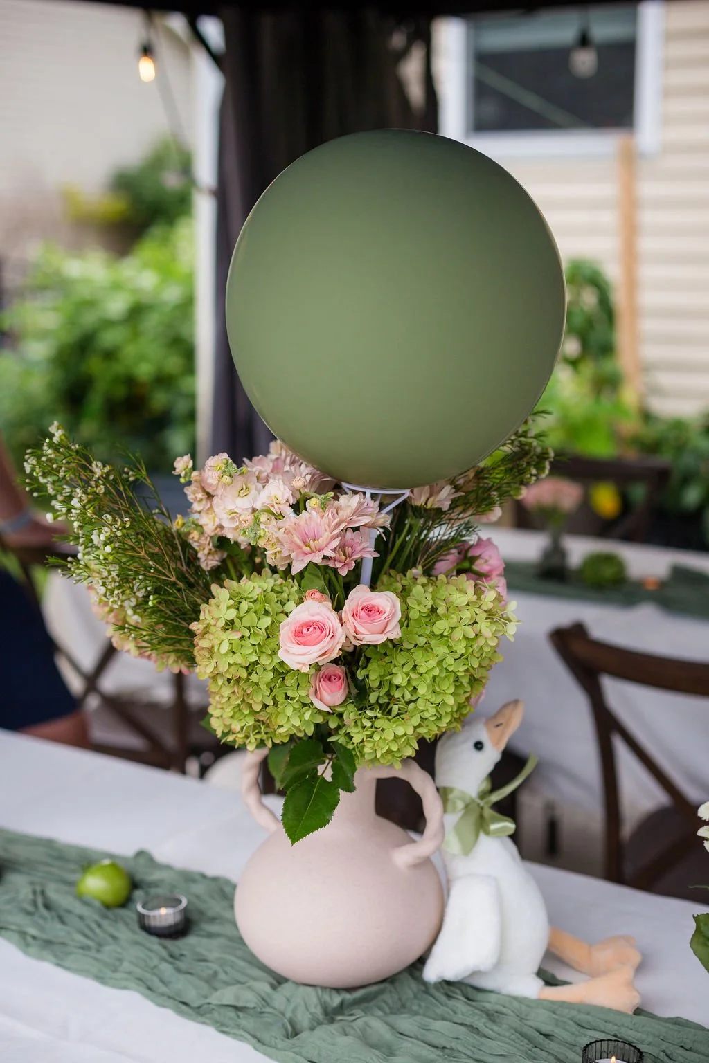 A table centerpiece with a pink ceramic vase filled with pink and green flowers, topped with a green balloon, and a plush toy swan sitting beside it, set on a green table runner.