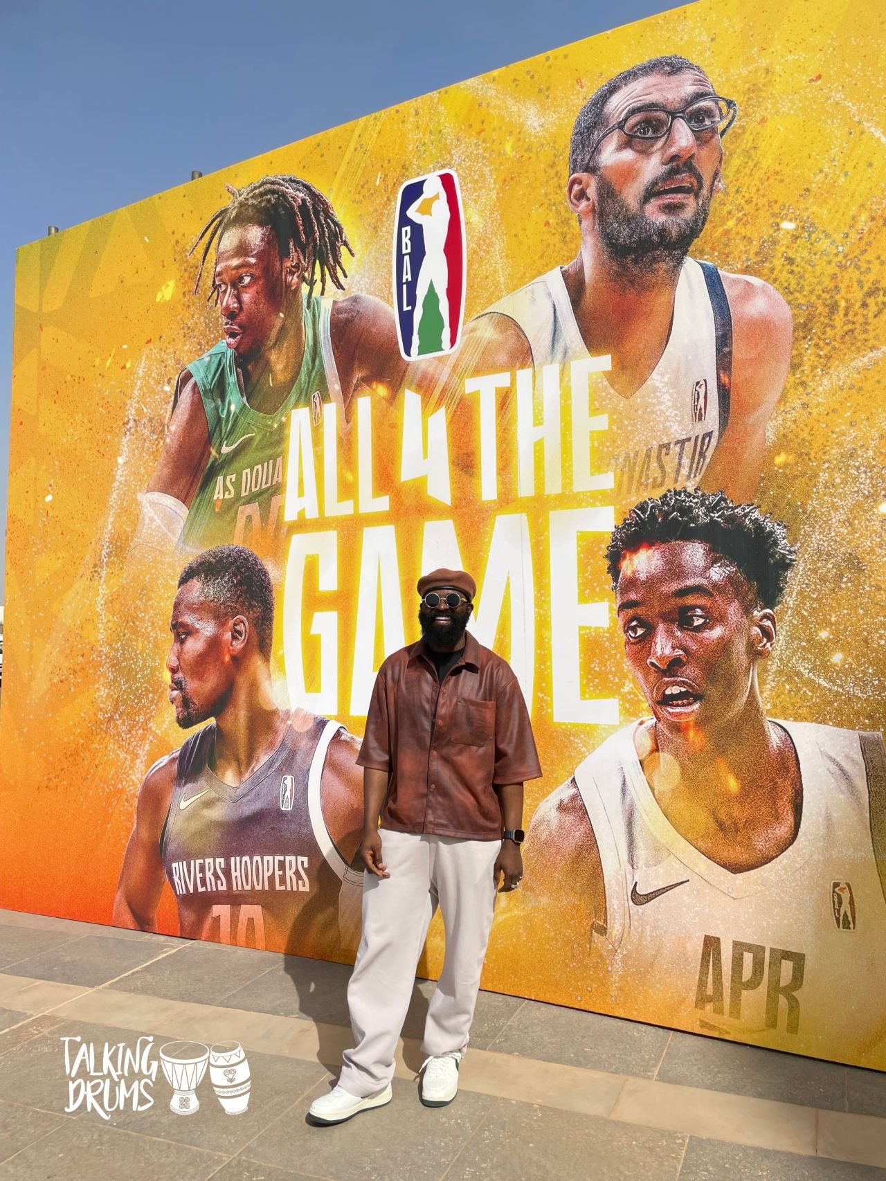 A person stands in front of a large colorful mural promoting the Basketball Africa League (BAL) with the text "All 4 The Game." The mural features portraits of basketball players from different teams such as AS Douane, Rivers Hoopers, and APR, each i