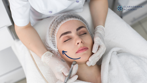 A woman with closed eyes receives a cosmetic treatment on her cheek from a professional wearing gloves, with the professional drawing a curved line on her face. There is a logo in the top right corner that says 'ClaraDerma Academy'.