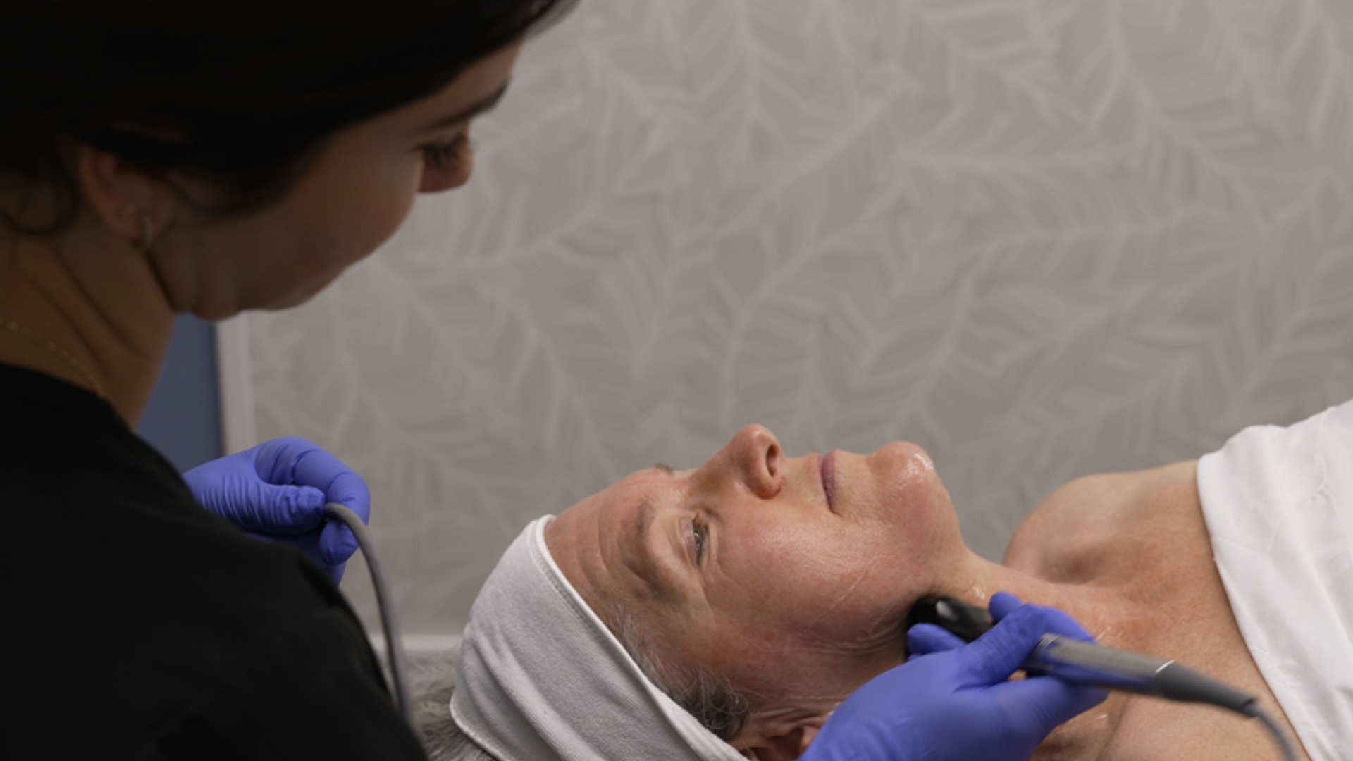 A woman receiving a facial treatment with a handheld device at a spa or clinic, with her eyes closed and wearing a white headband, lying on a treatment bed.