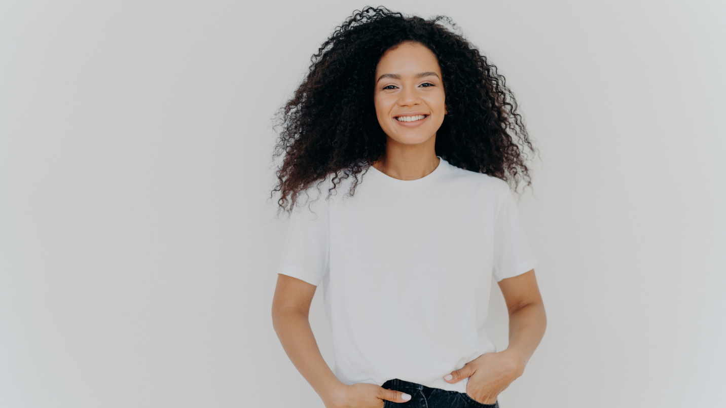 A young woman with curly hair smiling, wearing a white t-shirt and black pants, standing against a plain white background.