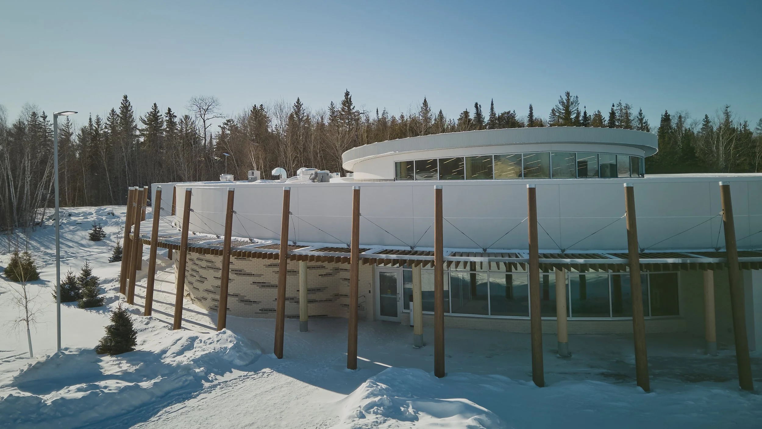 Modern building with curved roof and large glass windows, surrounded by snow-covered ground and evergreen trees in the background.