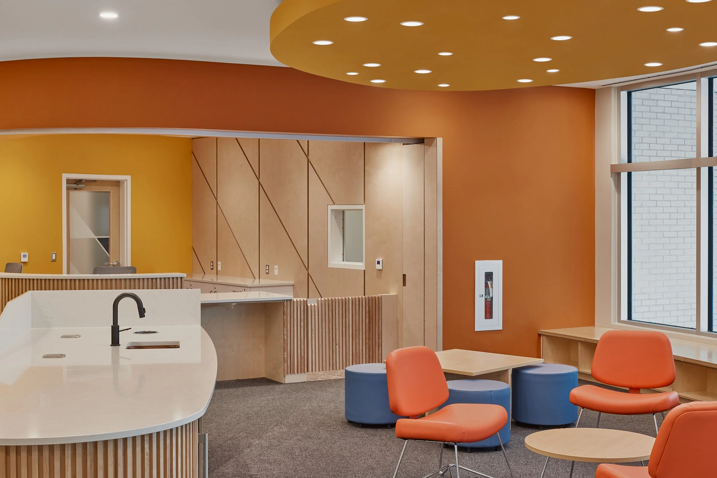 Modern waiting area with orange and blue chairs, a curved white counter with a sink, and colorful walls in yellow and orange, with large windows and a fire extinguisher on the wall.