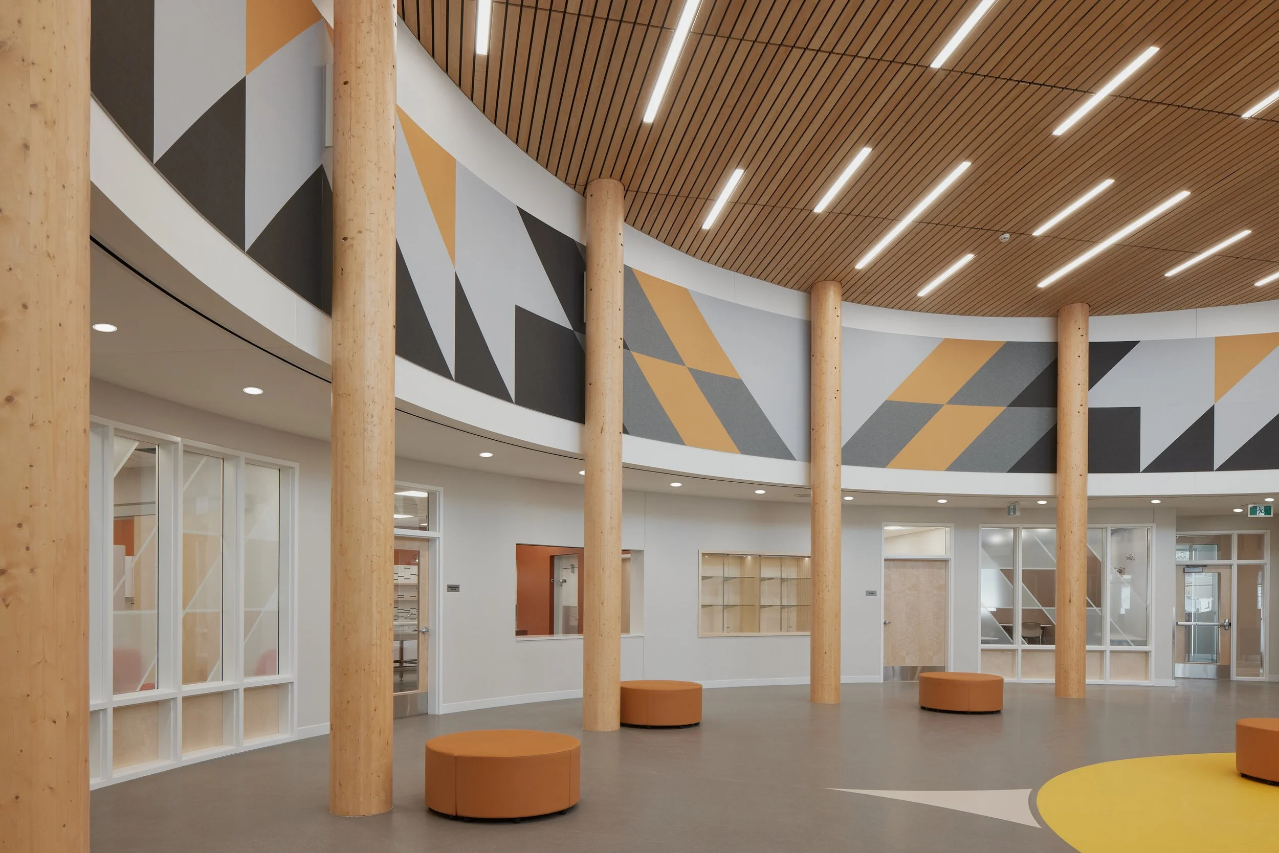 Modern interior space with wooden columns, geometric wall design in black, gray, and gold, wood-paneled ceiling with LED lighting, glass partitions, and orange seating.