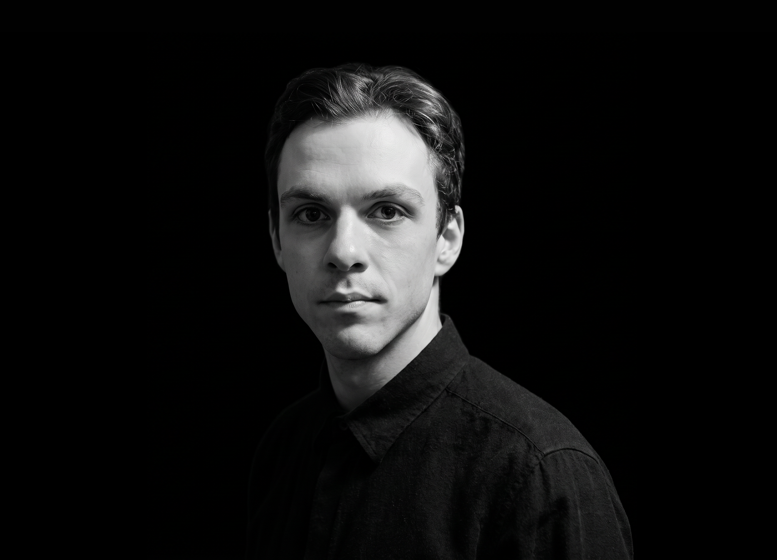 Andrew Falls, black and white portrait of a young man with short hair, wearing a dark shirt, against a solid black background.