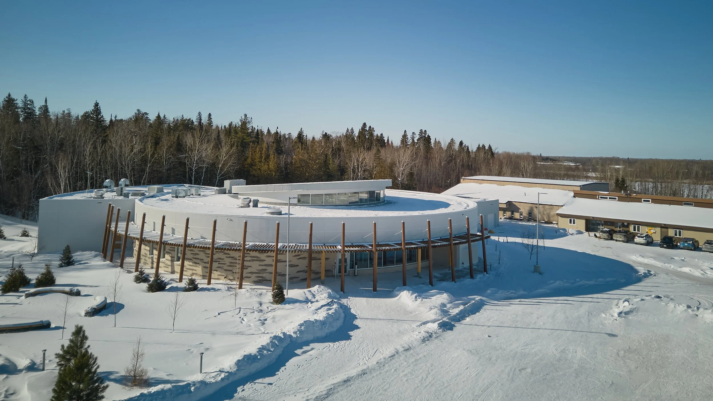 A modern, circular building with a flat roof covered in snow, surrounded by snow-covered landscape and trees, with a parking lot and additional buildings nearby under a clear blue sky.