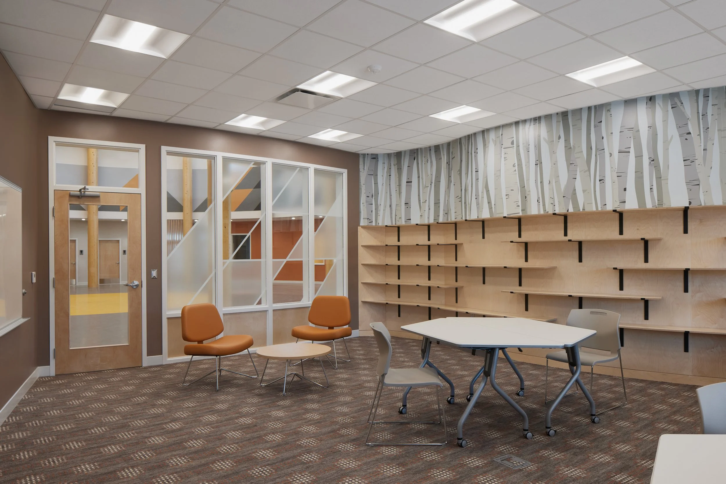 Empty modern office or community room with chairs, table, wooden shelving, patterned wall, glass dividers, and ceiling lights.