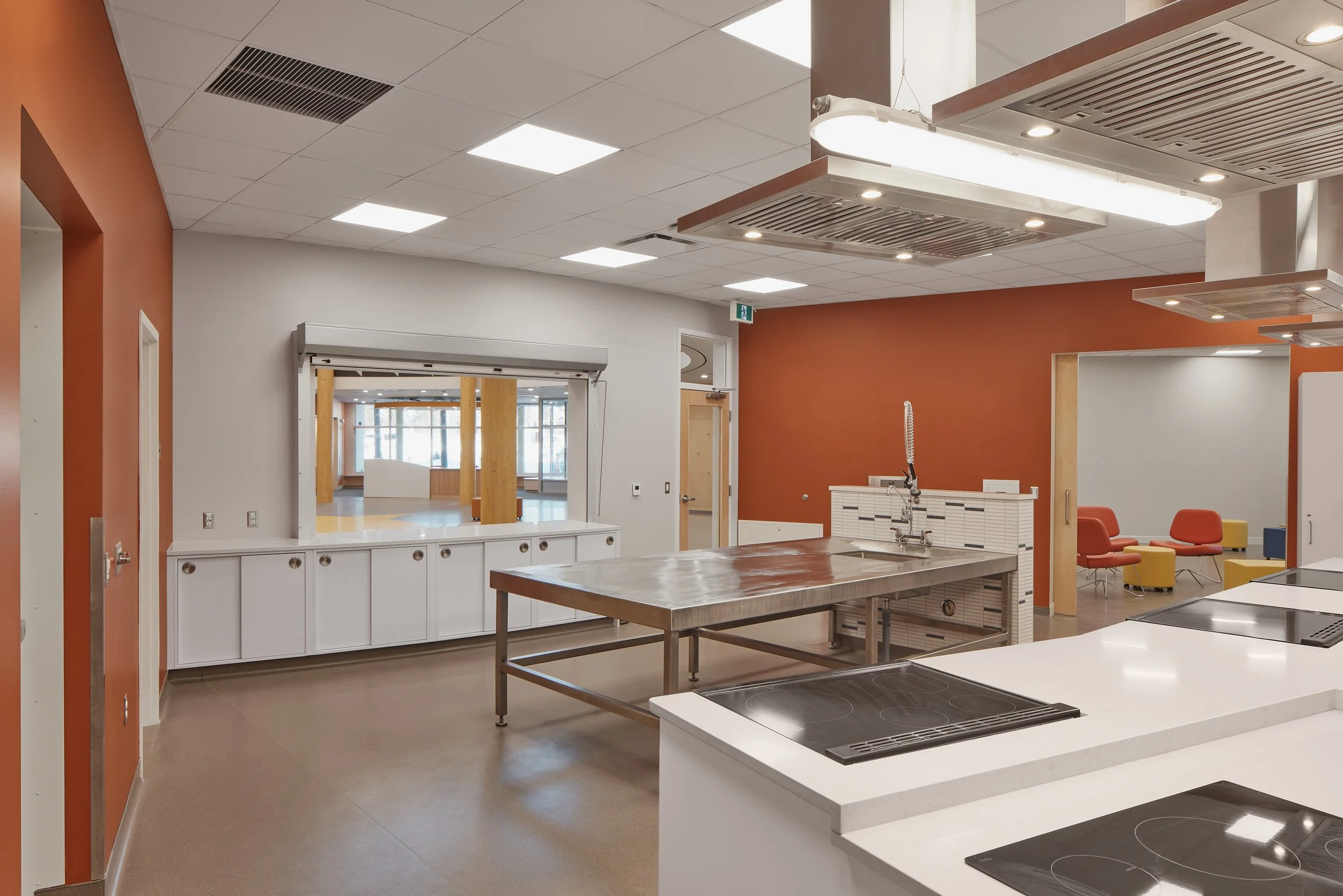 Modern kitchen with stainless steel island, white countertops, and orange and white walls. Seating area with colorful chairs visible in the background.