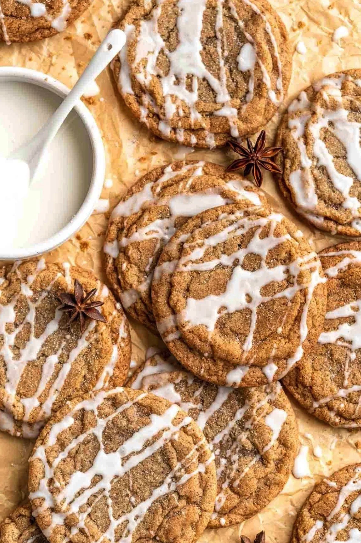 Chocolate crinkle cookies drizzled with white icing, with a small cup of milk on a wooden surface, decorated with star anise.