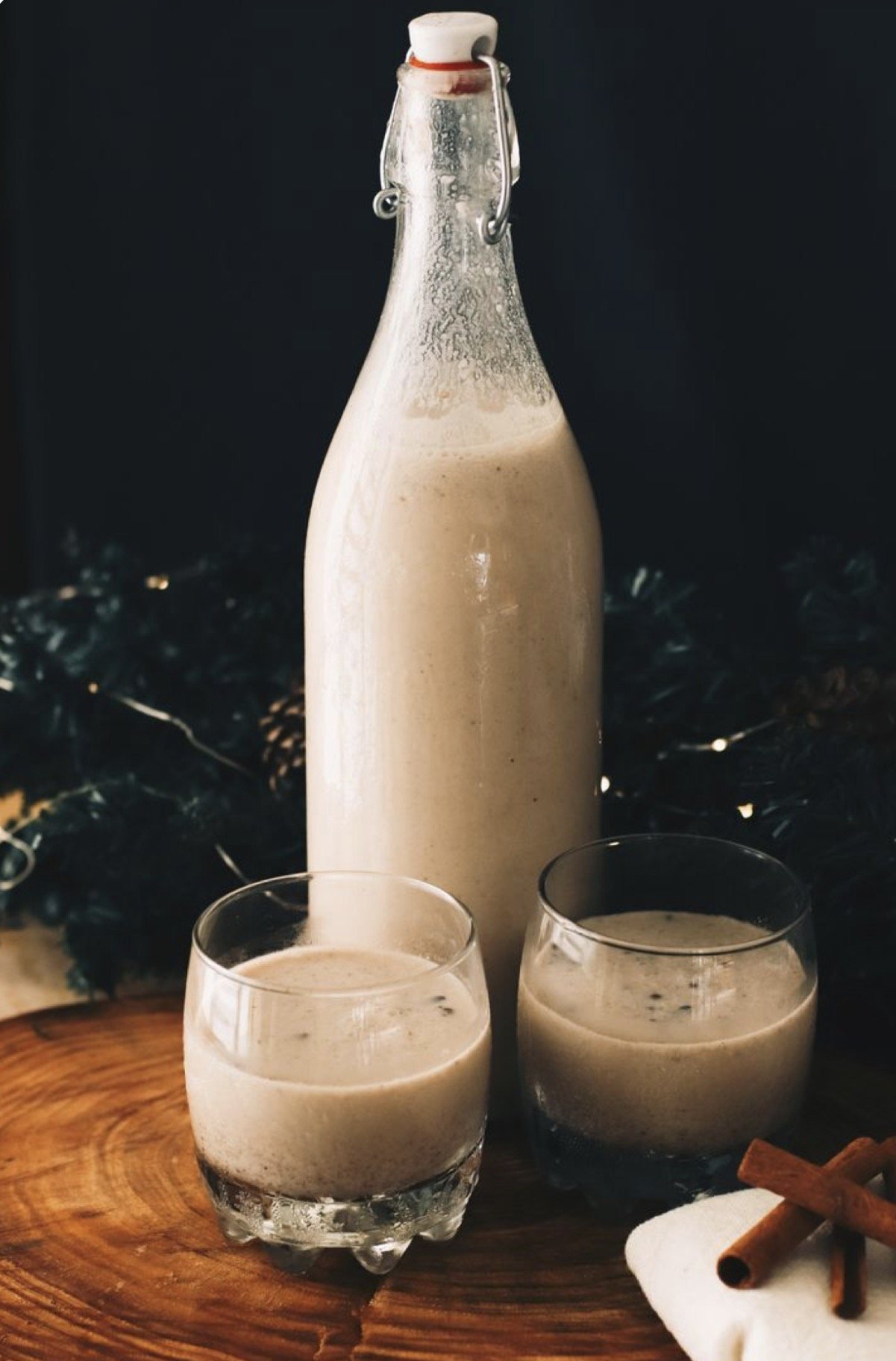 Glass bottle filled with a creamy, light brown beverage, two short glasses of the same drink, and cinnamon sticks on a white napkin on a wooden surface with a dark background.