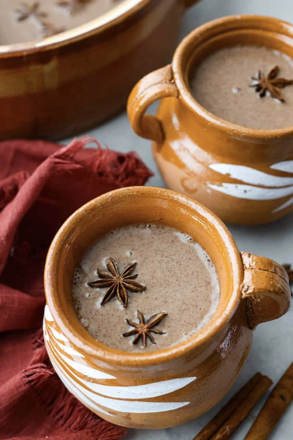 Two ceramic mugs of hot chocolate topped with star anise, placed on a table with a red cloth and cinnamon sticks.
