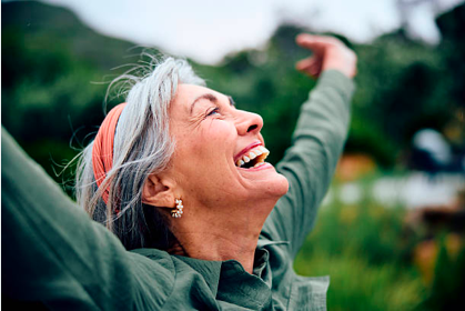 An elderly woman with gray hair and an orange headband, smiling and raising her arms outdoors in a green, natural setting.