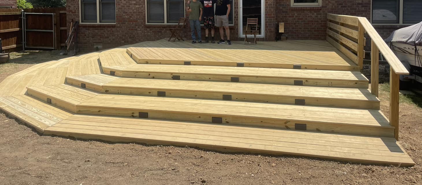 Freshly constructed wooden deck with multiple levels and steps, situated in a backyard with a brick house, outdoor furniture, and a boat in the background.