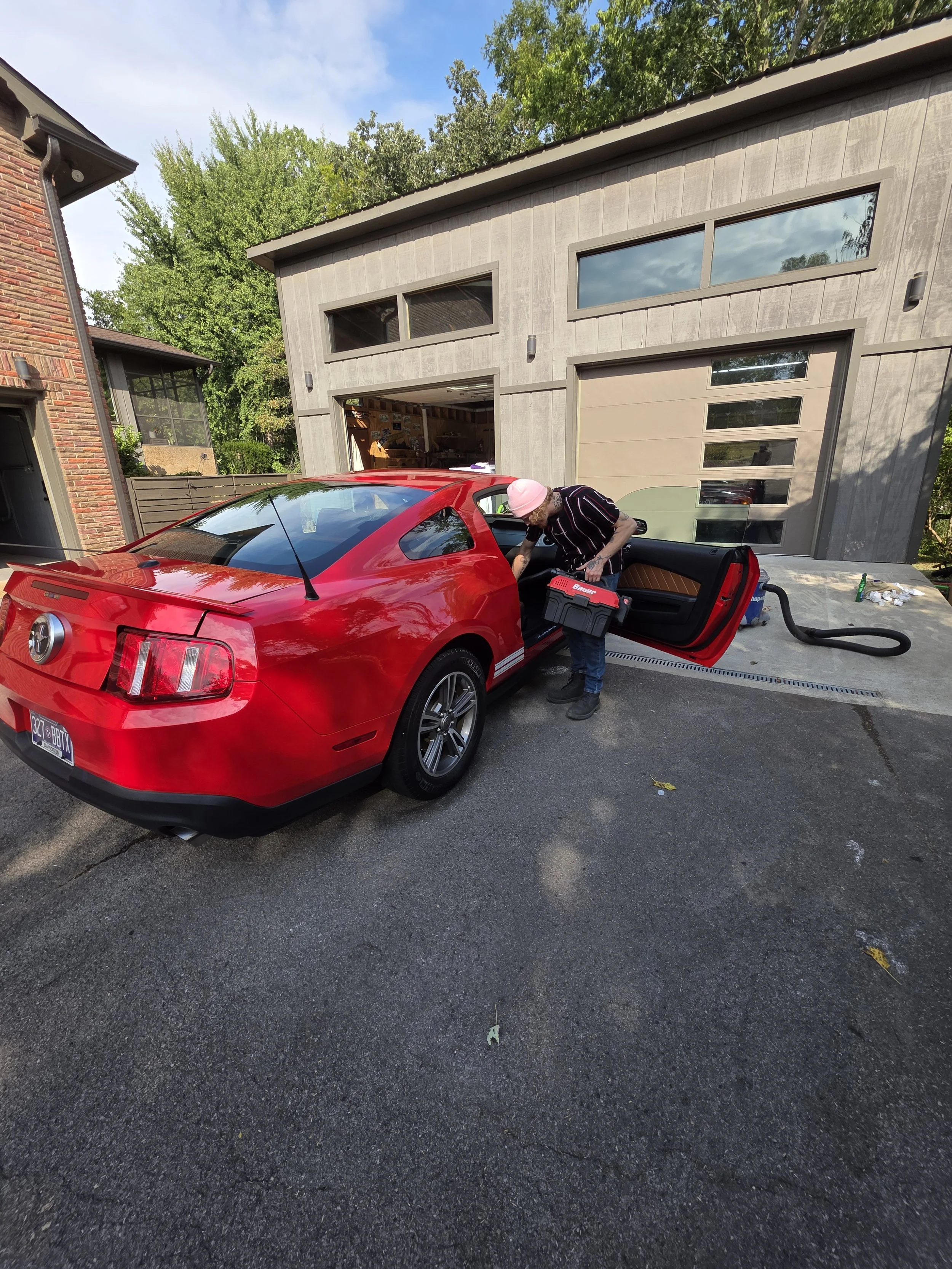 A person with a pink head wrap and striped shirt is using a shop vacuum to clean a red Ford Mustang parked in a driveway in front of a modern house with a garage.