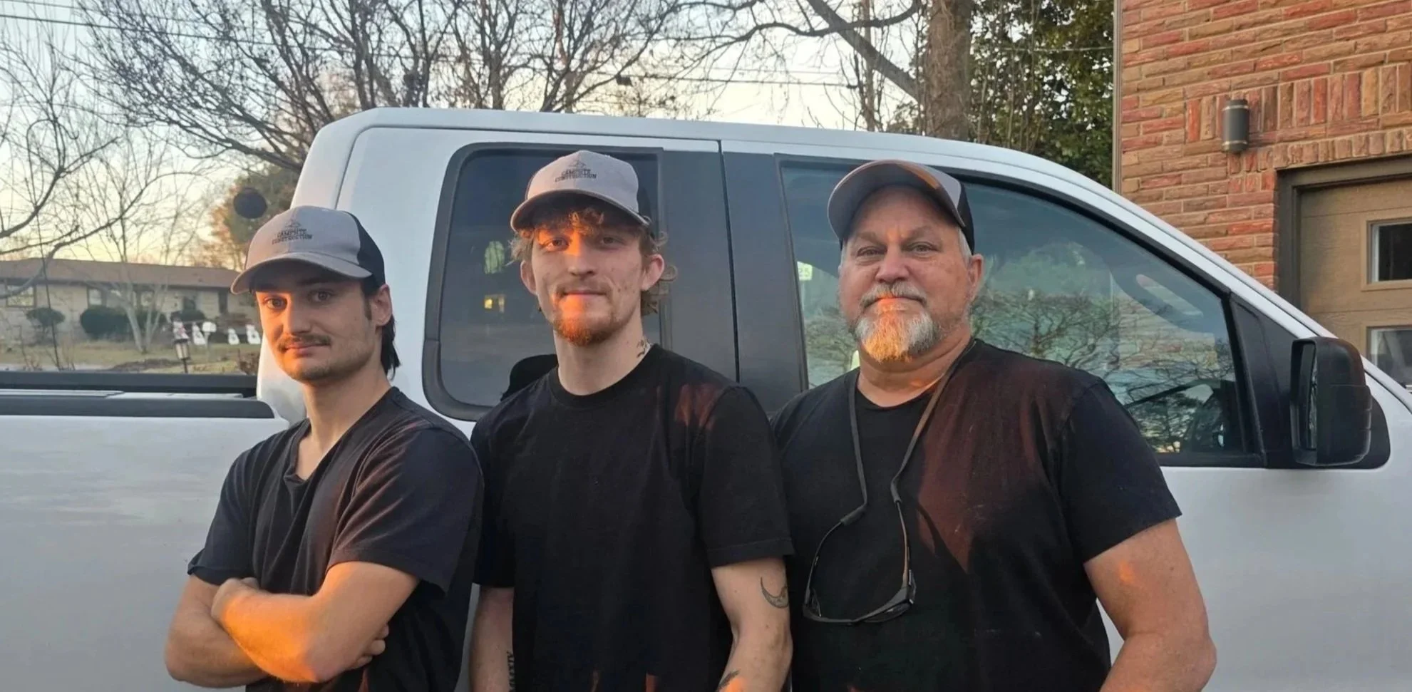 Three men standing side by side outdoors near a white vehicle with trees and a brick house in the background during sunset.