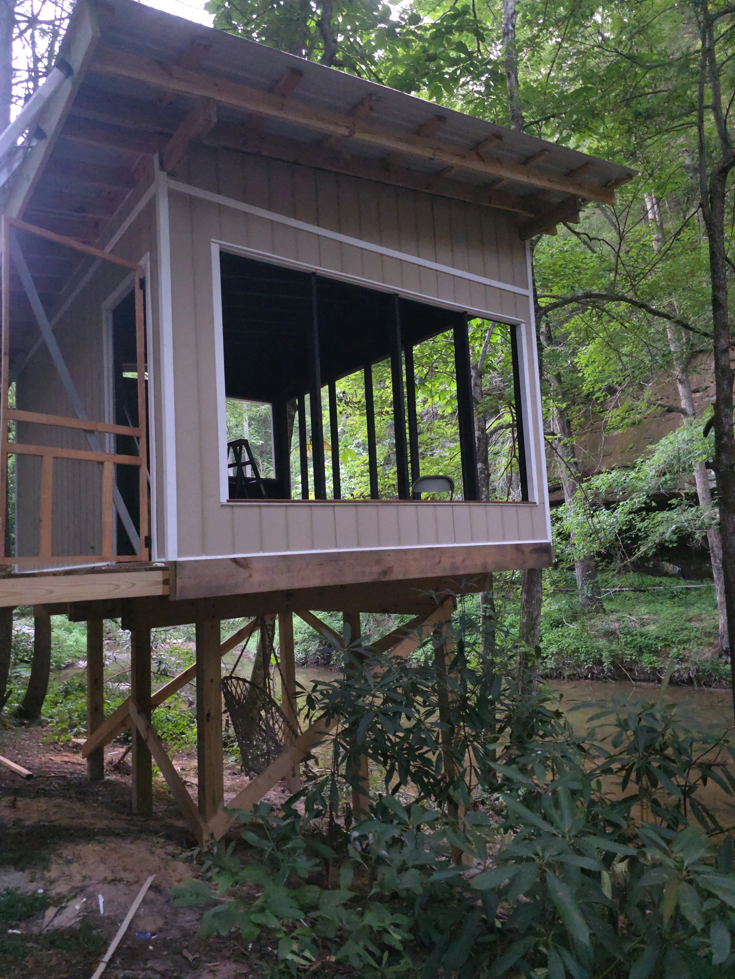 A small unfinished cabin or treehouse on stilts in a wooded area, with visible interior framing and window openings, surrounded by trees and foliage.