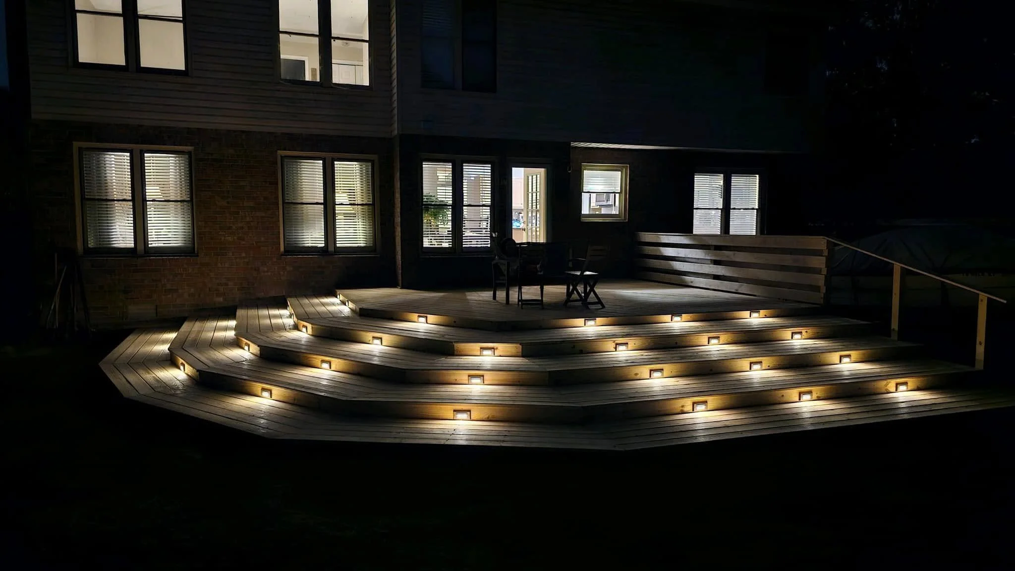 Night view of a wooden deck with stairs leading up to it, illuminated by embedded ground lights, attached to a house with multiple windows, some with blinds, and an outdoor seating area with chairs and a table.