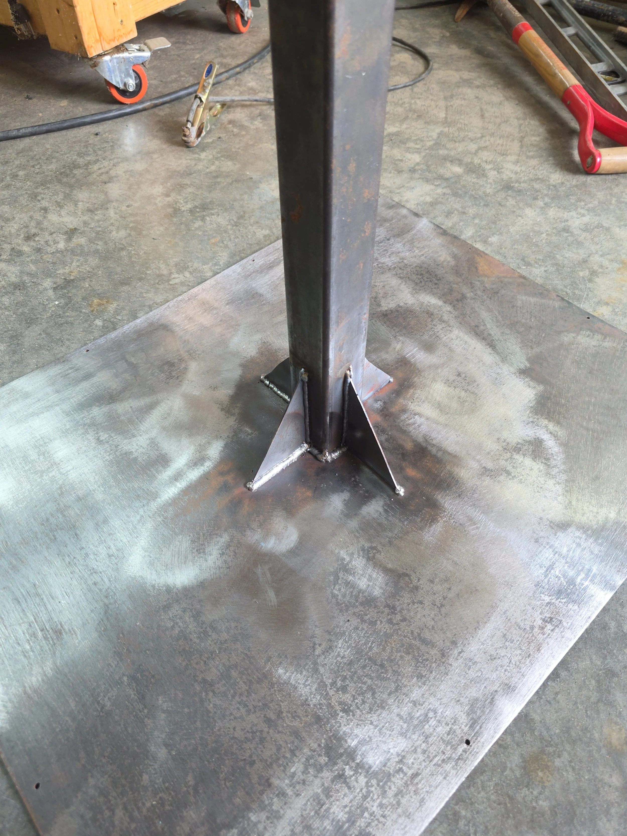 Steel pole welded to a flat metal base plate with triangular gussets at the bottom of the pole for added support, on a workshop floor.