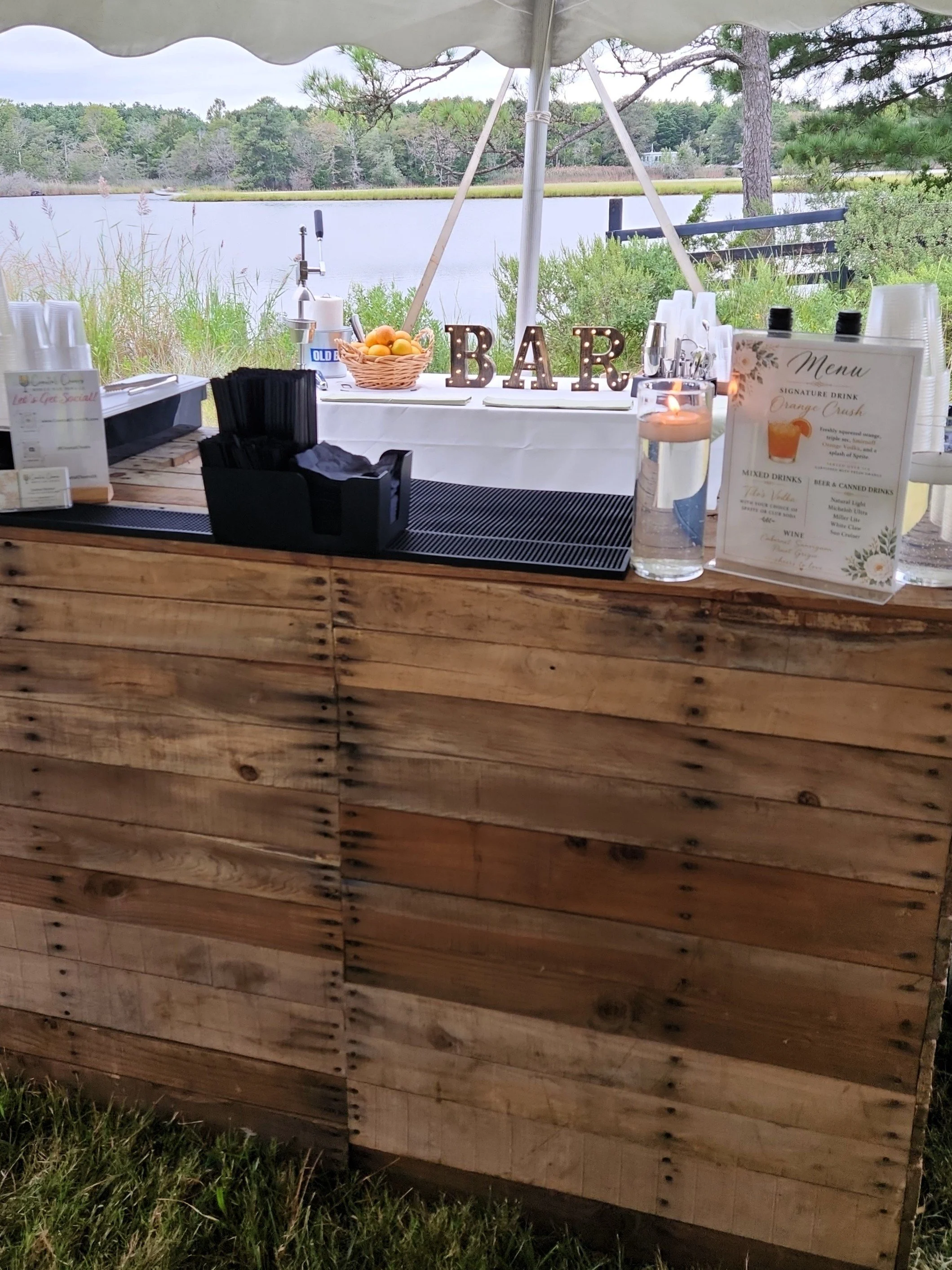 A lakeside outdoor bar setup under a canopy with decorative 'BAR' letters, a basket of lemons, a water tap, and a menu stand on a wooden counter, overlooking a pond with trees and grass in the background.
