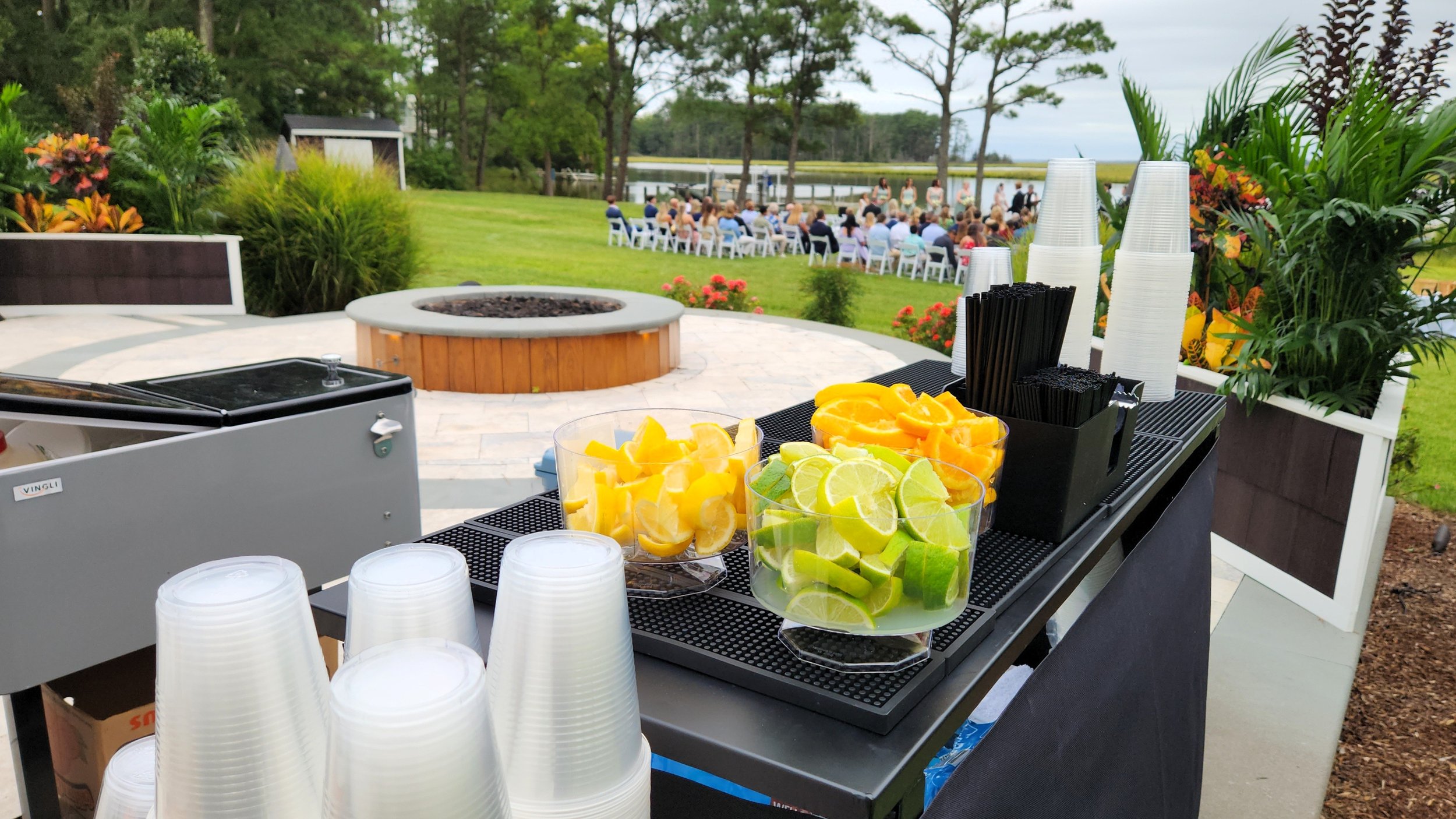 Conference or event outdoors with a refreshment table in the foreground containing cups, sliced citrus and fruit in clear bowls, with guests seated on chairs facing a stage or speaker near a body of water in the background.