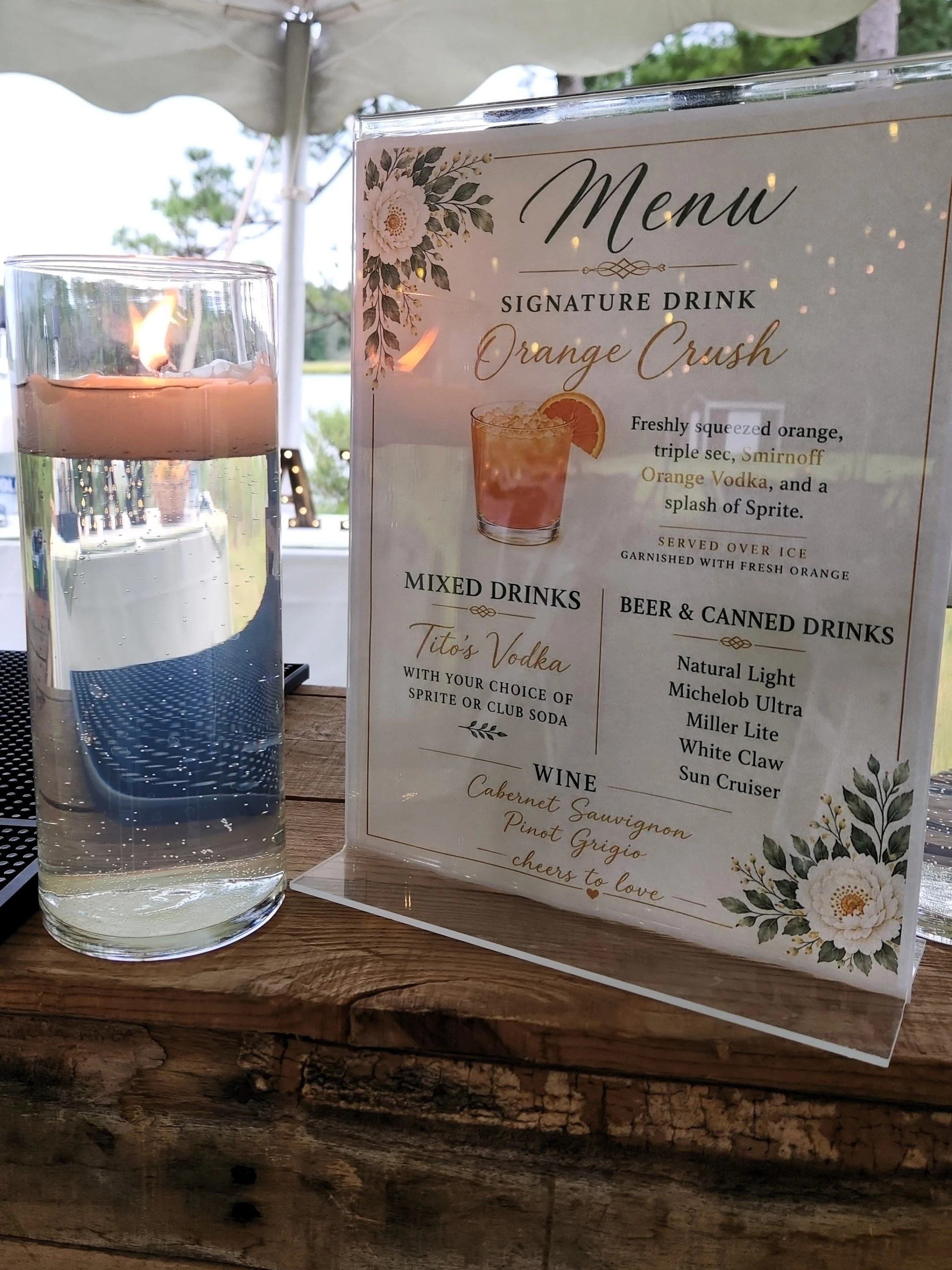 A menu card and a candle placed on a wooden table at an outdoor setting. The menu features a signature drink called Orange Crush, which includes orange, triple sec, Smirnoff Orange Vodka, and Sprite. The menu also lists mixed drinks, beer, and wine o