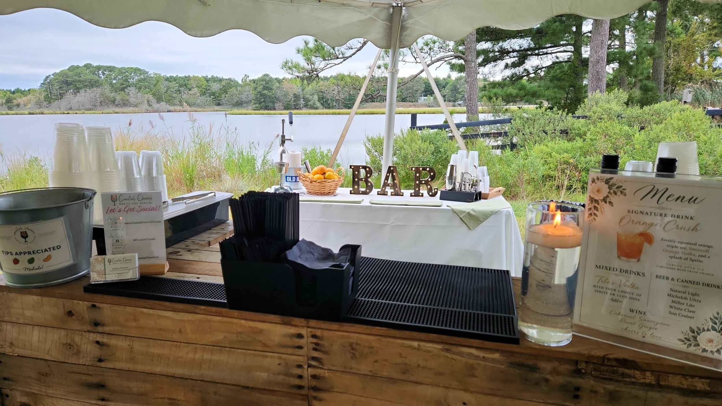 A lakeside outdoor bar setup with cups, a basket of oranges, a lit candle, and a menu, under a canopy with trees and water in the background.
