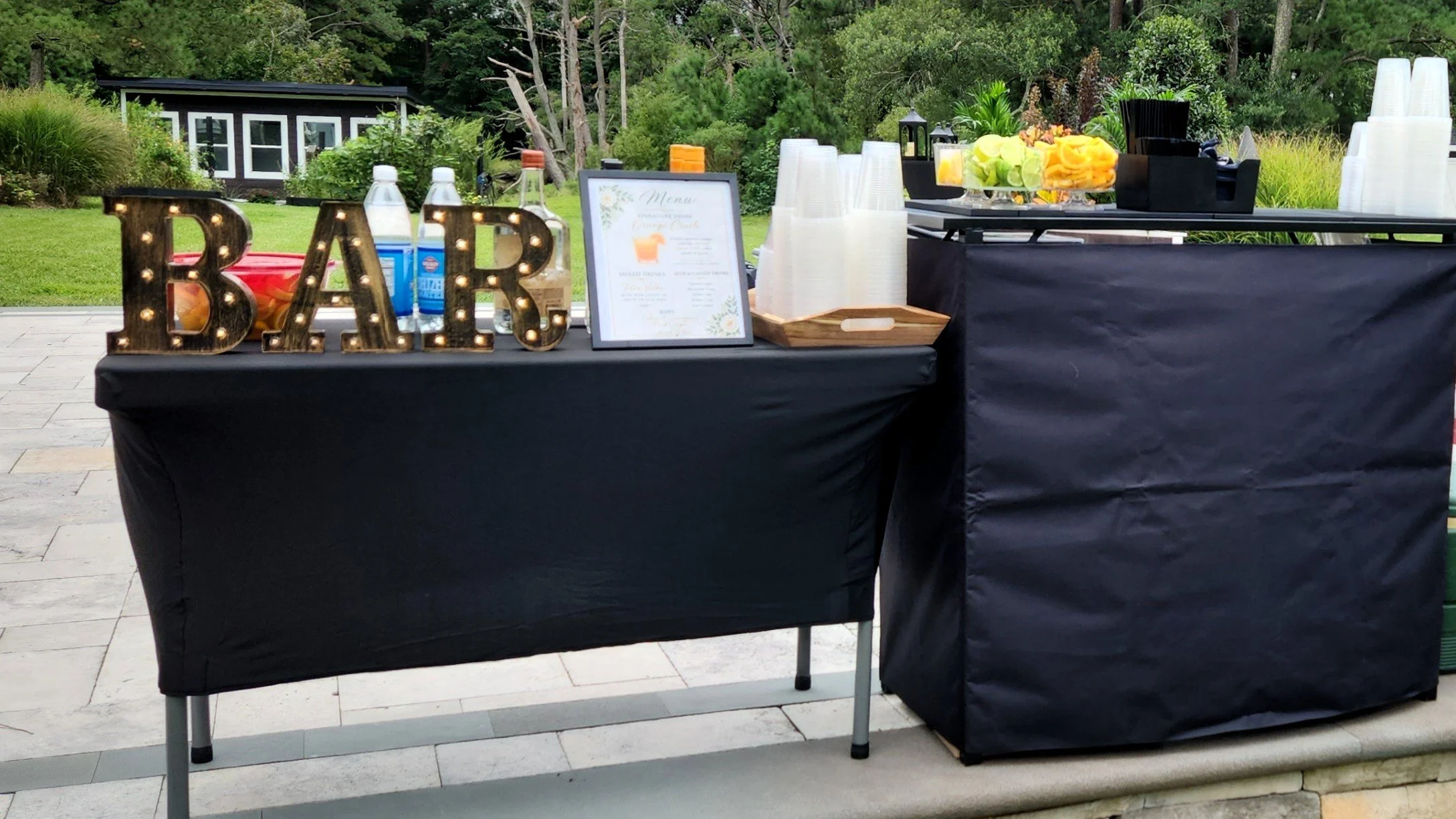 Outdoor party buffet table with black tablecloth, large illuminated "BAR" sign, menu frame, water bottles, plastic cups, flowers, and serving items, set against a lush green backyard.