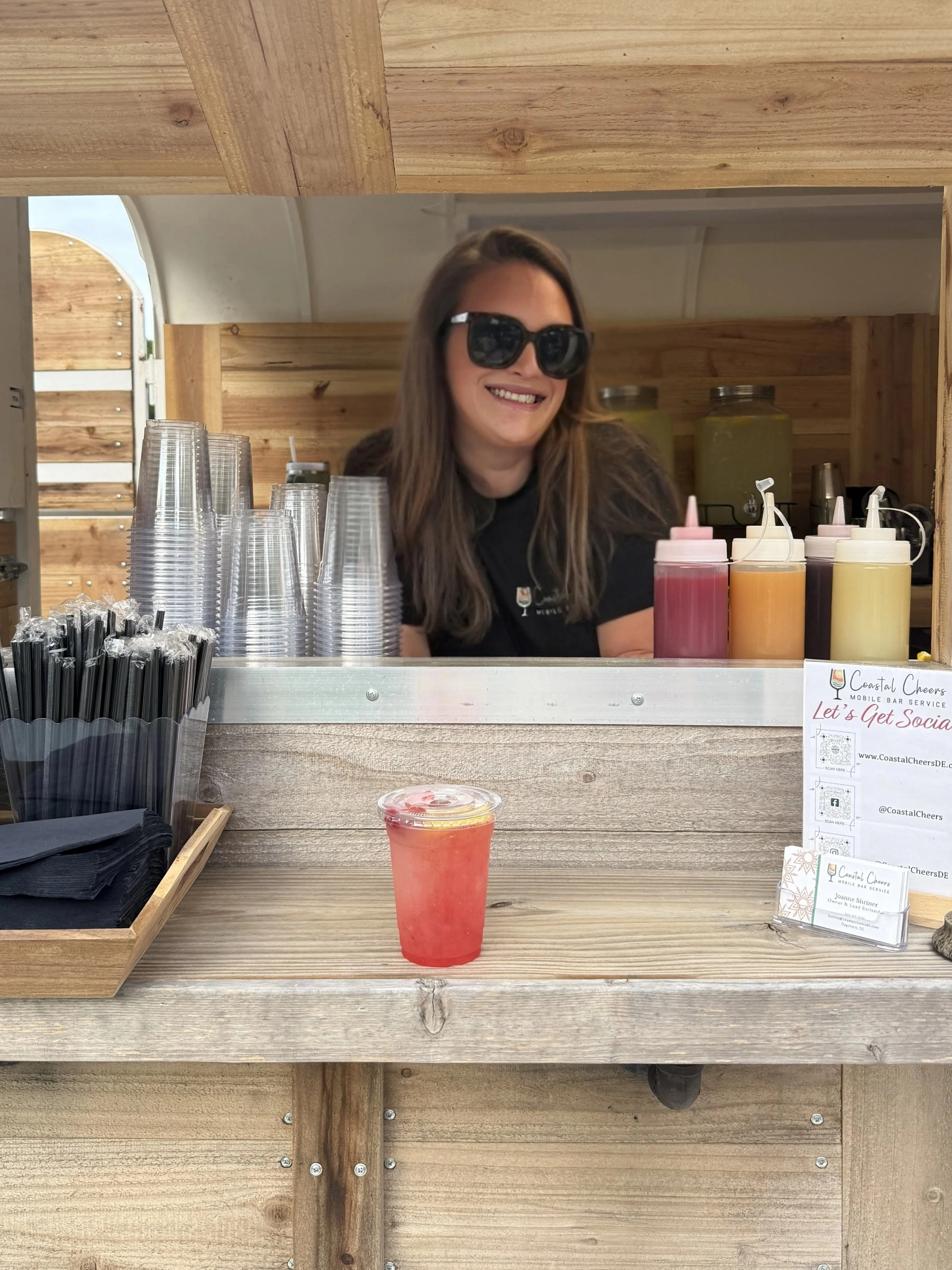 A woman with long brown hair, wearing black sunglasses, is smiling behind a bar counter with colorful squeeze bottles of drink flavorings and a clear plastic cup with a pink-colored beverage in front of her. The bar has a wooden background and a sign