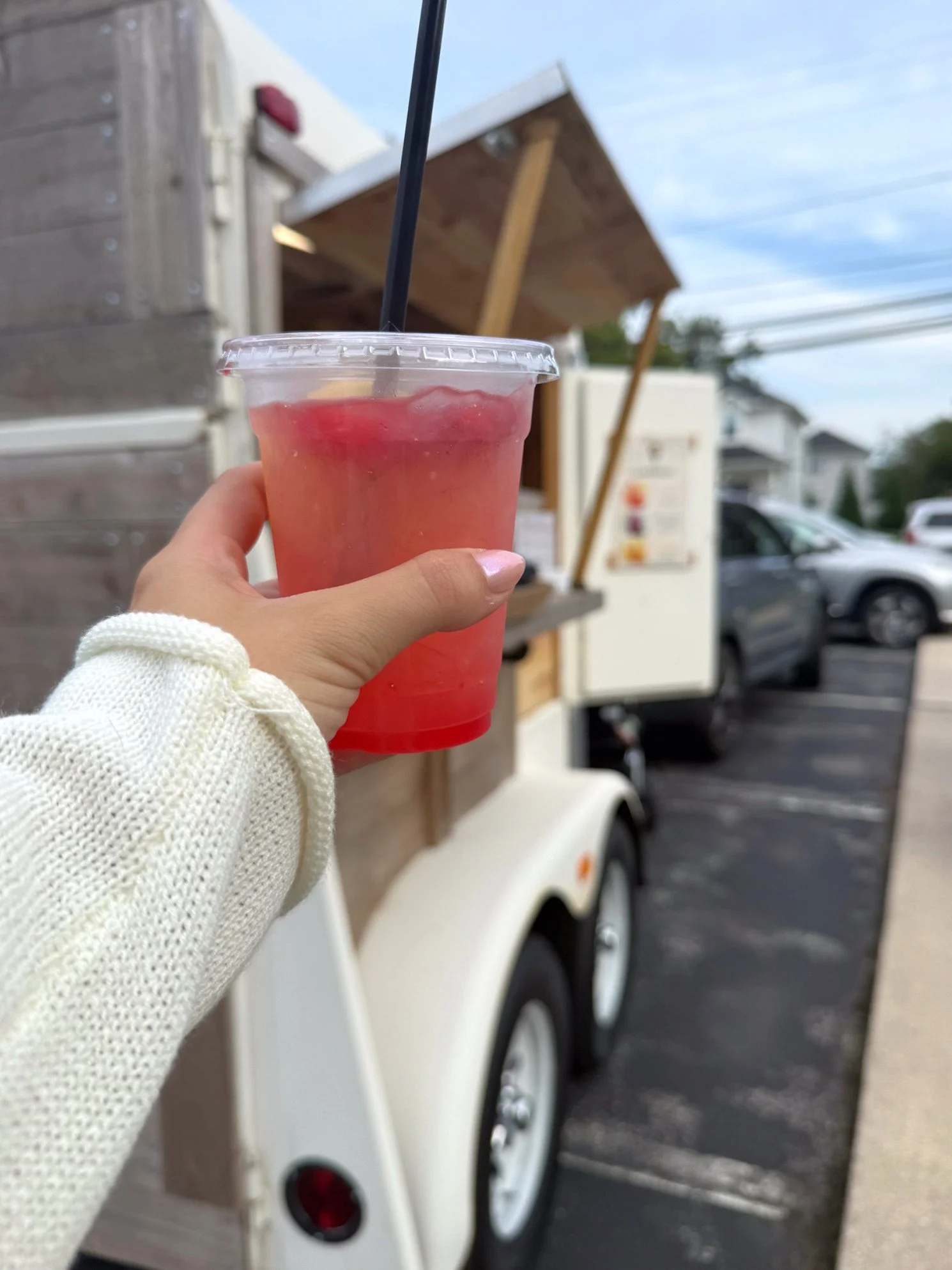 A hand holding a pink beverage in a clear plastic cup with a black straw, with a food truck in the background.