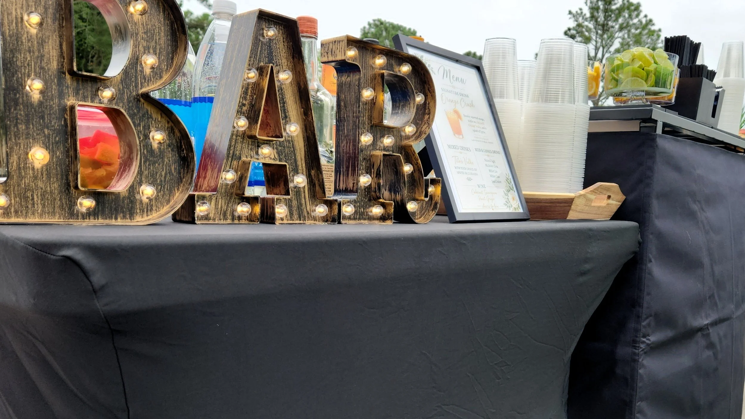 Decorative table display with large illuminated "BAR" sign, framed menu, and beverage station including clear cups and containers with limes, lemons, and black straws, set outdoors.