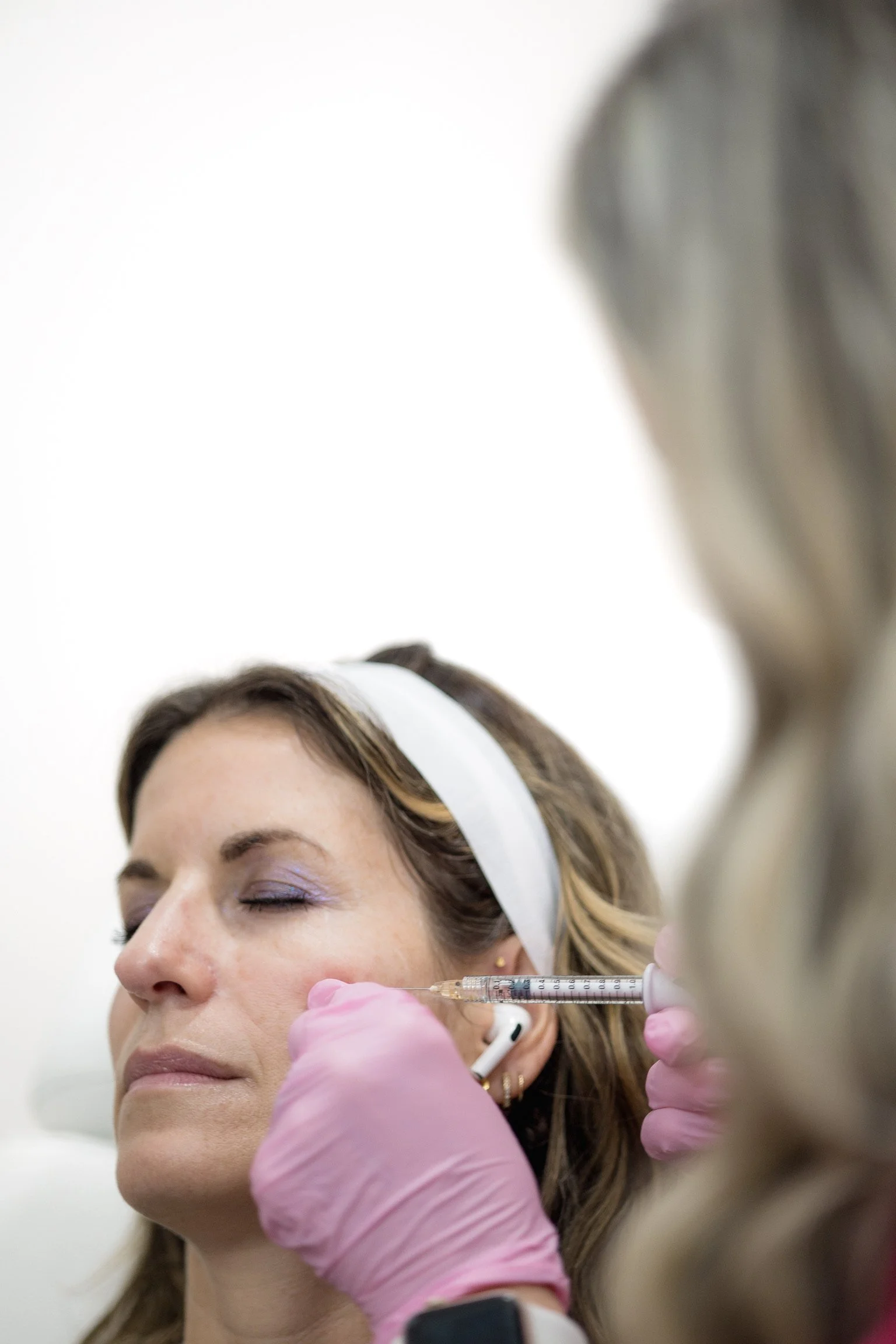 A woman with a headband receiving a cosmetic injection in her cheek from a healthcare professional wearing pink gloves.