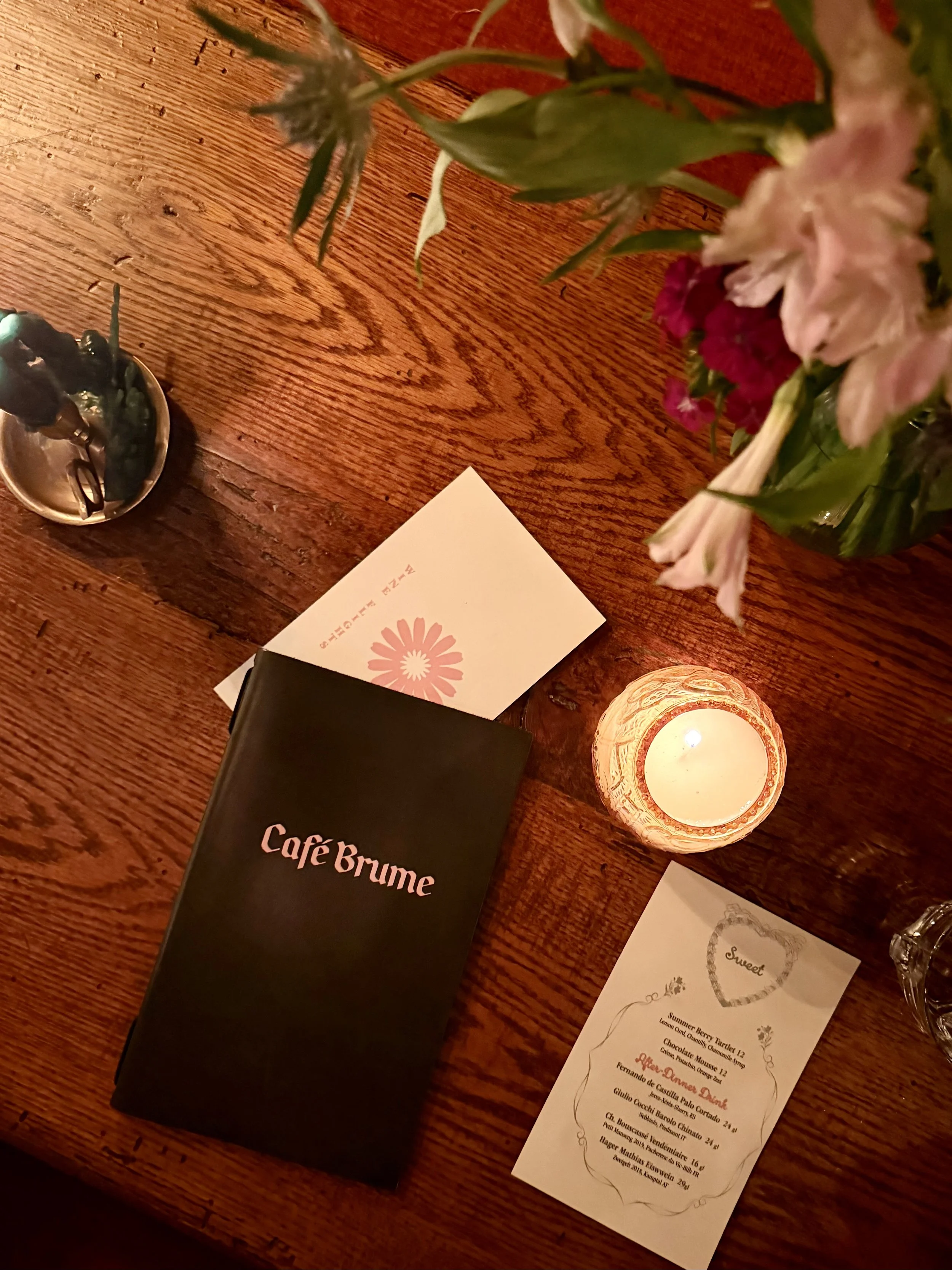 A wooden table with a black menu titled 'Café Brume,' a candle in a decorative holder, a bouquet of flowers in a vase, and a printed menu or flyer with a heart-shaped design and text.