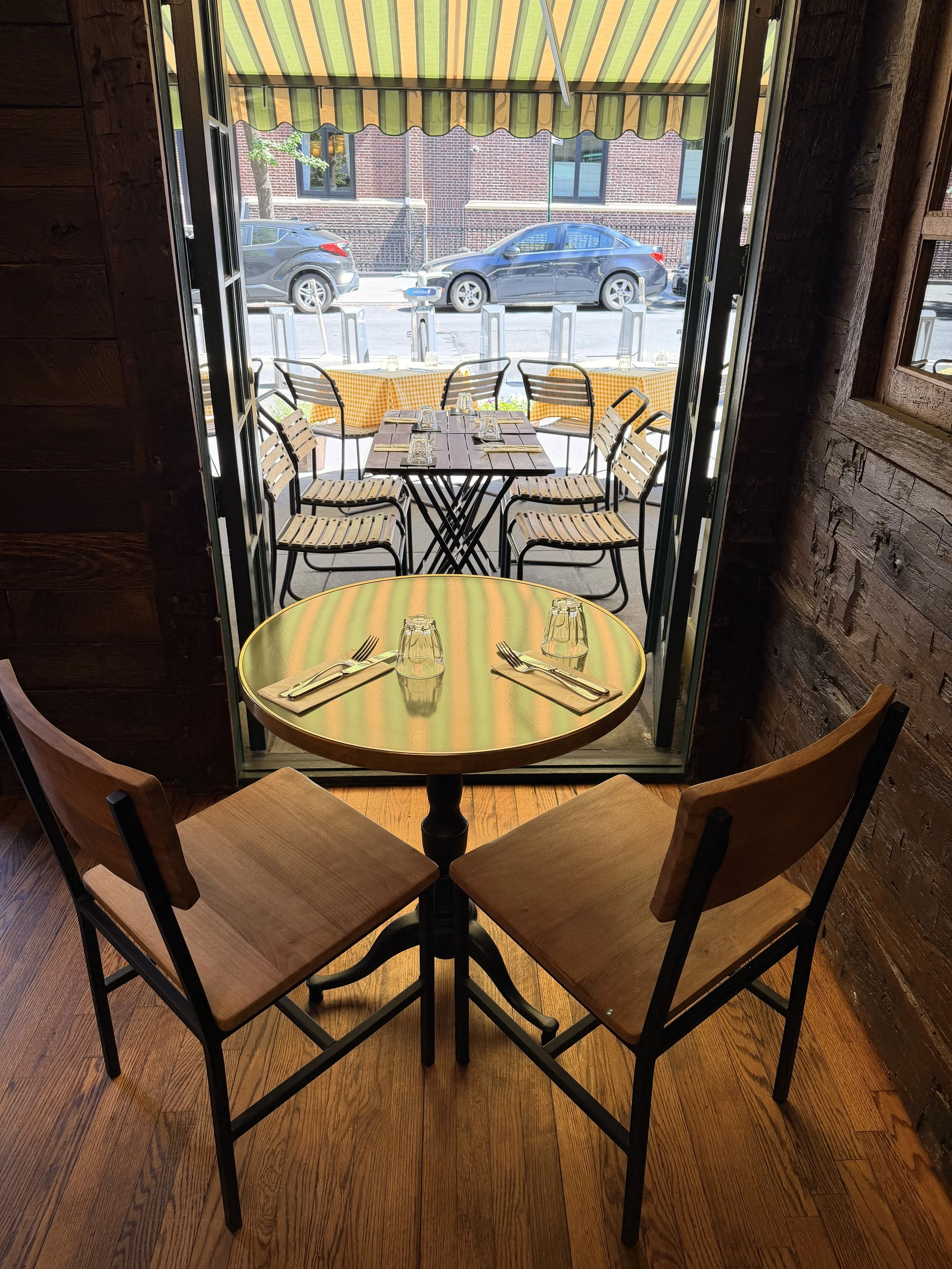 Inside view of a restaurant or cafe with a round table set for two, plates, glasses, and silverware. Outside view through an open door.